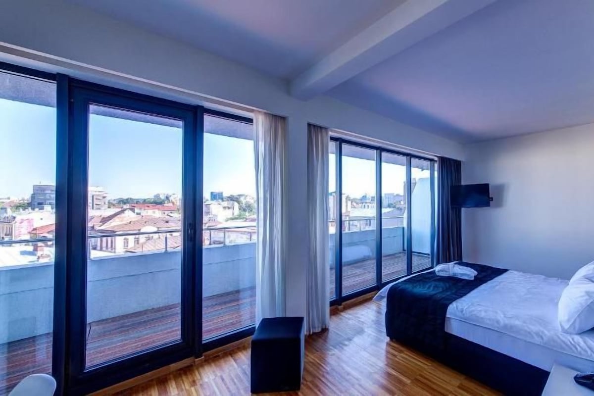 Quarto de hotel com uma cama grande, televisão e vista da cidade através de amplas janelas.