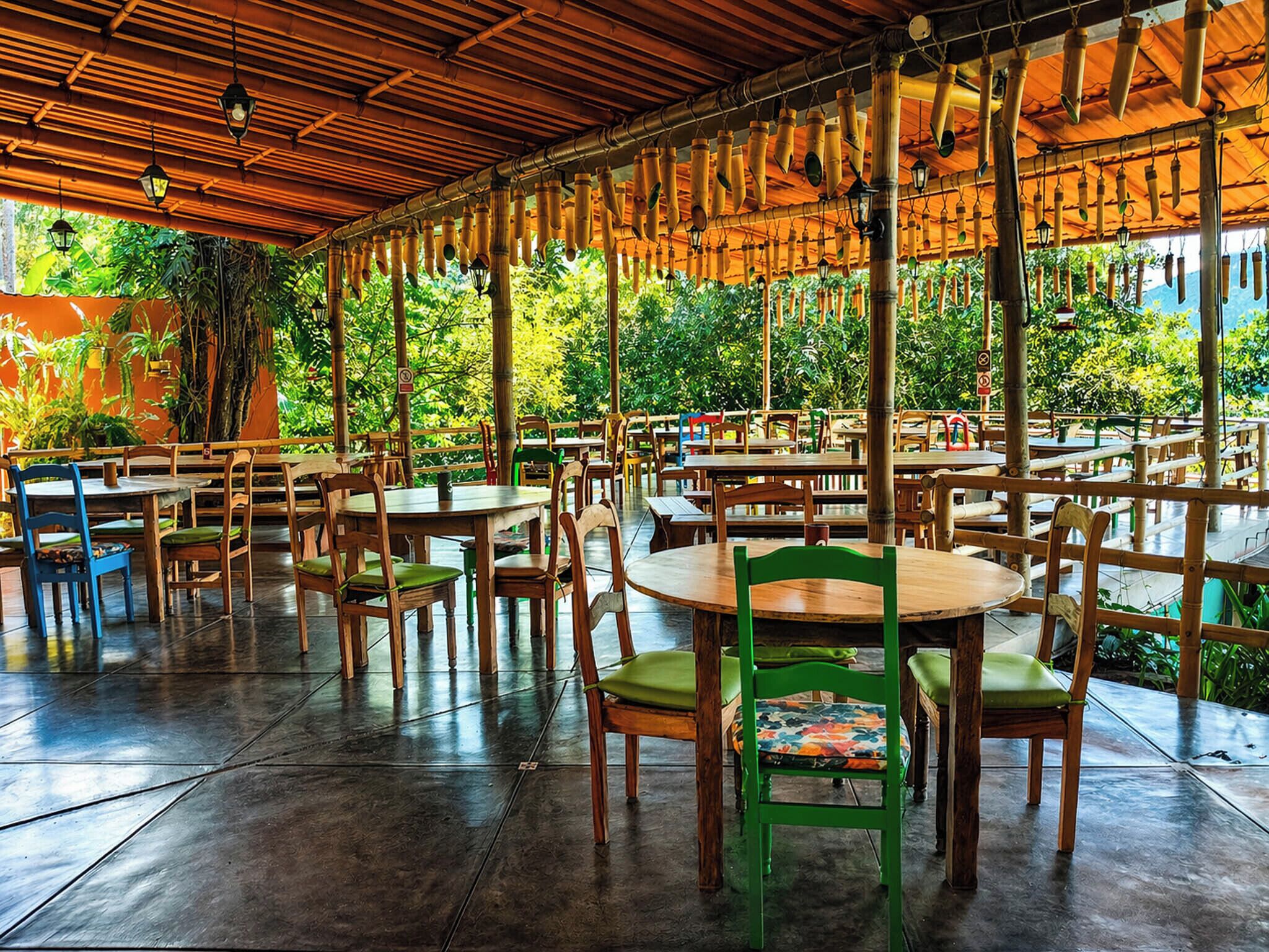 An outdoor dining area with tables and chairs, surrounded by greenery and a bamboo ceiling.