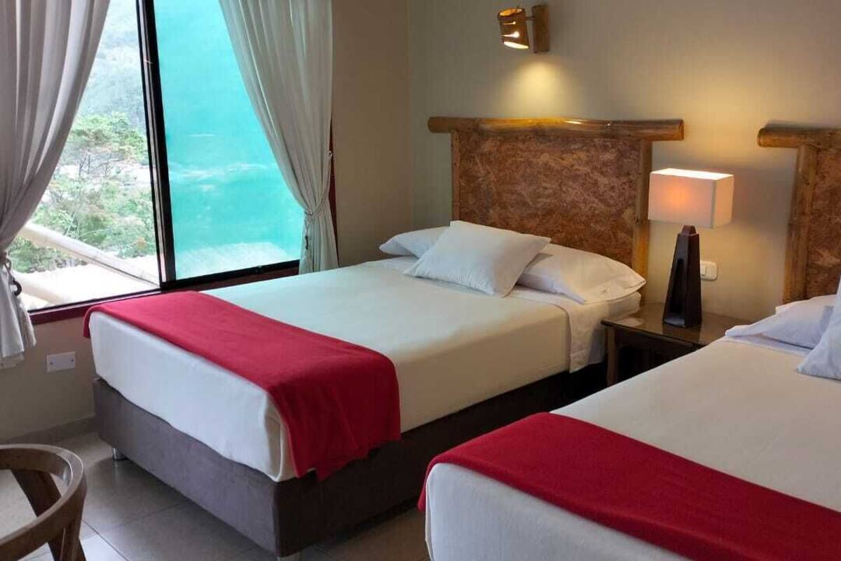 Habitación de hotel con dos camas, cada una con sábanas blancas y colchas rojas. Hay una ventana con cortinas, una mesita de noche con lámpara y una silla.