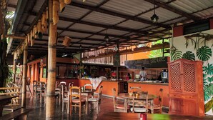 Un restaurante con columnas de bambú, mesas y sillas de madera, una barra con un menú y un mural de plantas tropicales.
