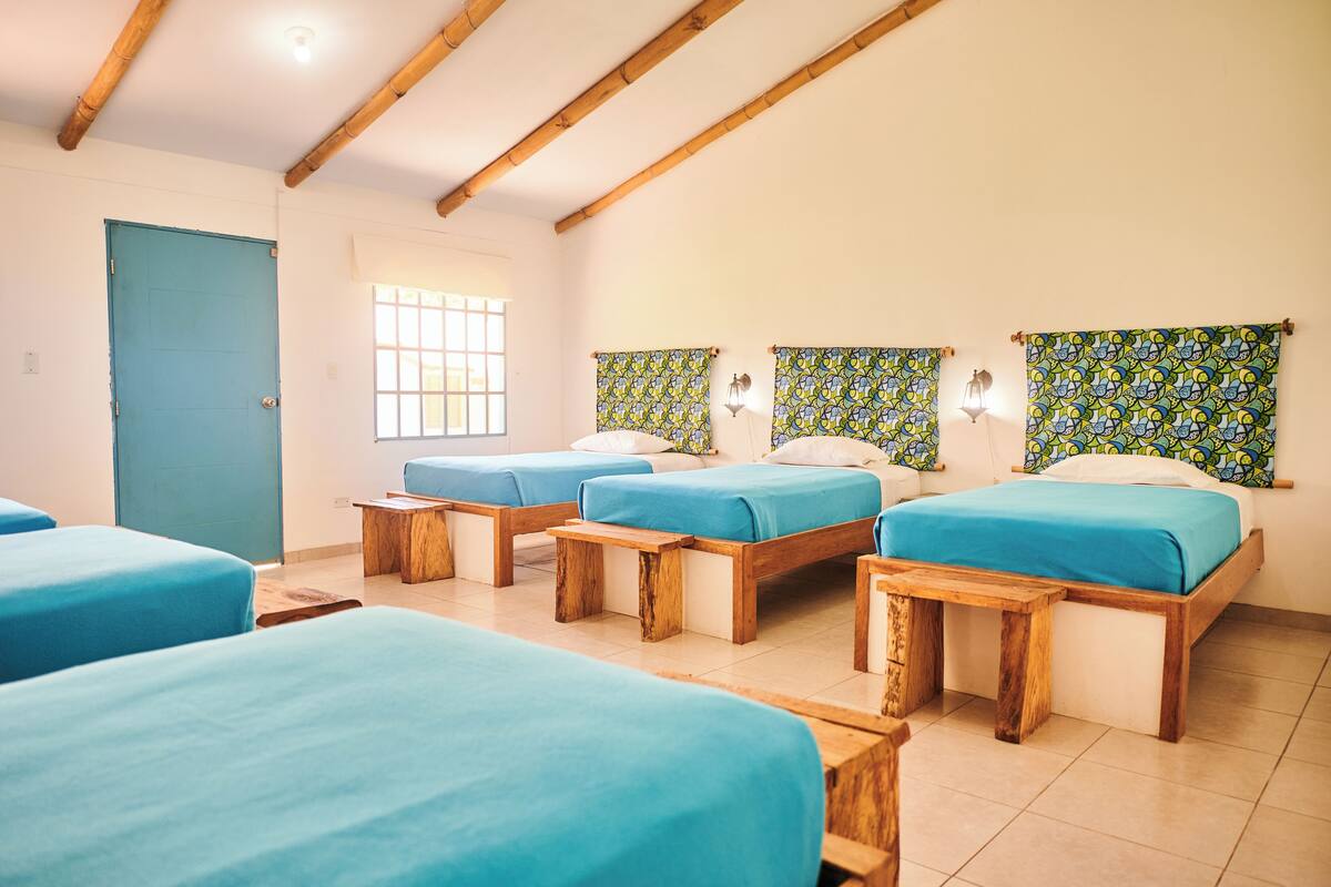 Una habitación con cuatro camas, cada una con estructura de madera y ropa de cama azul. Hay bancos de madera al pie de cada cama. El piso es de baldosas y el techo de madera presenta vigas a la vista.