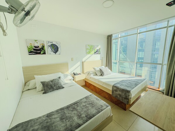 Habitación de hotel con dos camas, una mesita de noche, un ventilador y amplias ventanas con vistas a la ciudad.