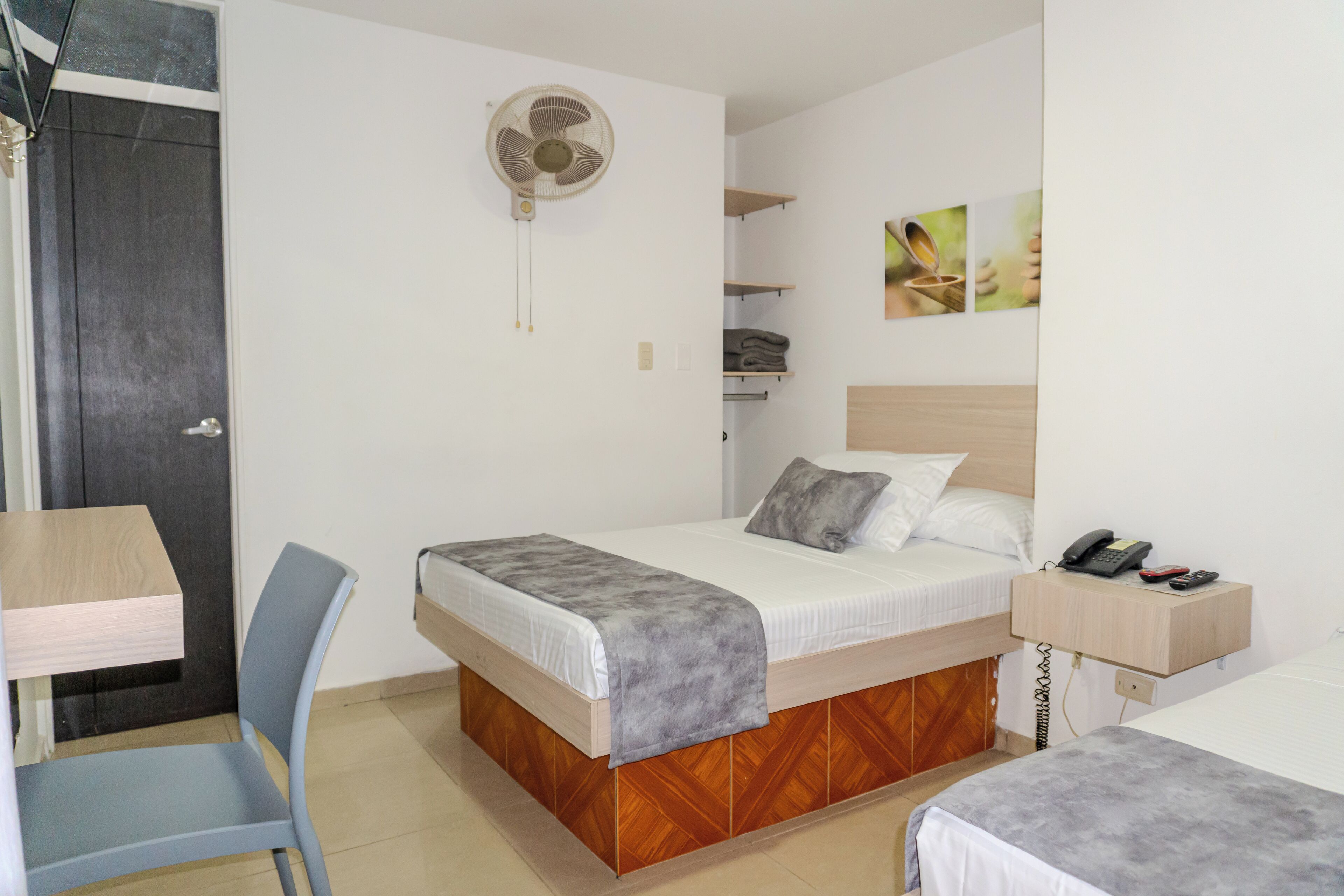 Una habitación de hotel con cama, una silla, un escritorio, un ventilador, un teléfono y obras de arte en la pared.