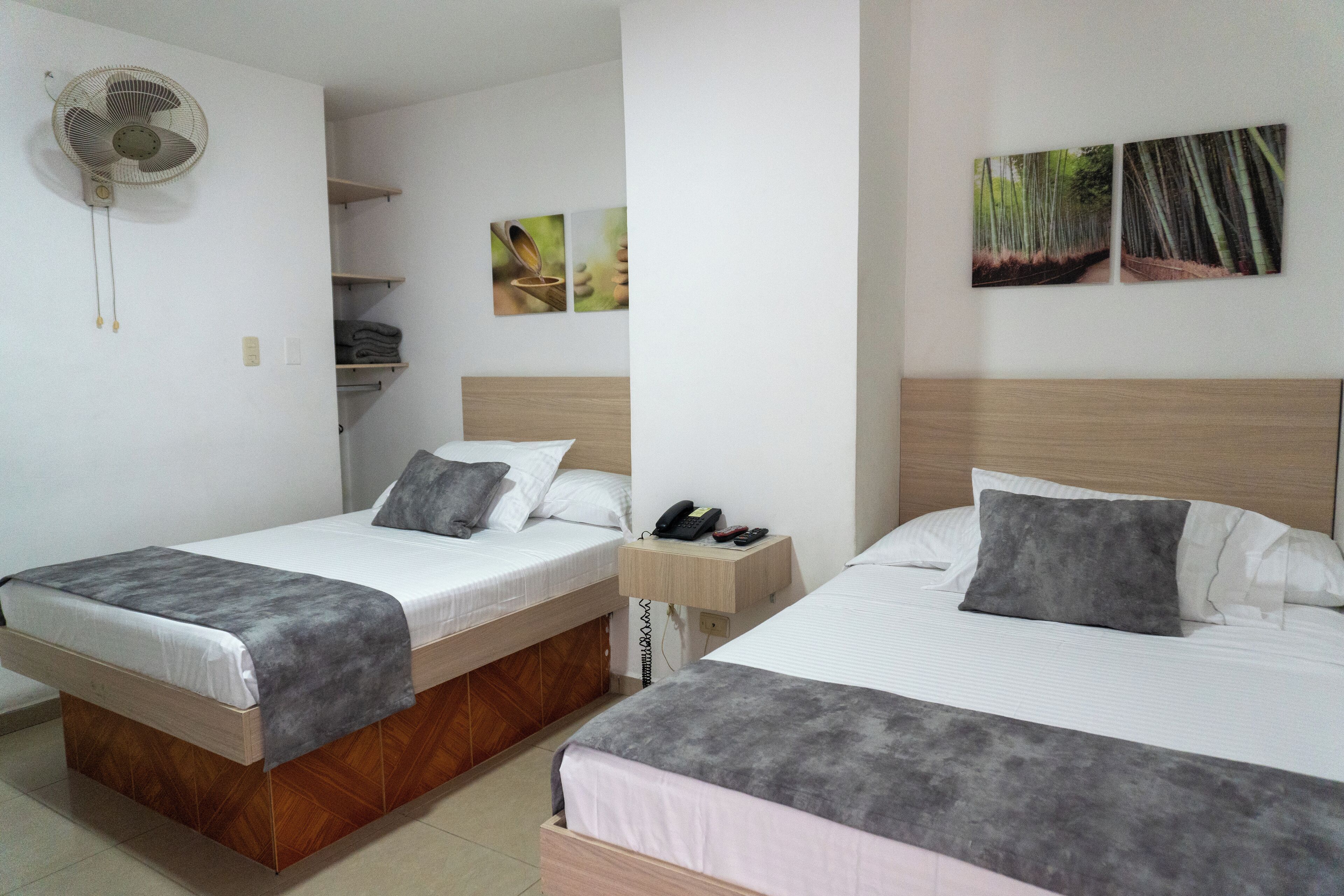 Une chambre d’hôtel avec deux lits, une table de chevet avec un téléphone et des clés, un ventilateur mural et des œuvres d’art représentant des forêts de bambous.