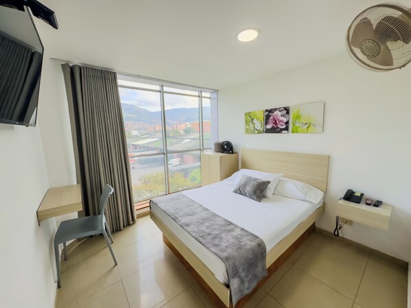 A hotel room with a bed, a desk, a chair, a TV, a fan, and a window with a city view.