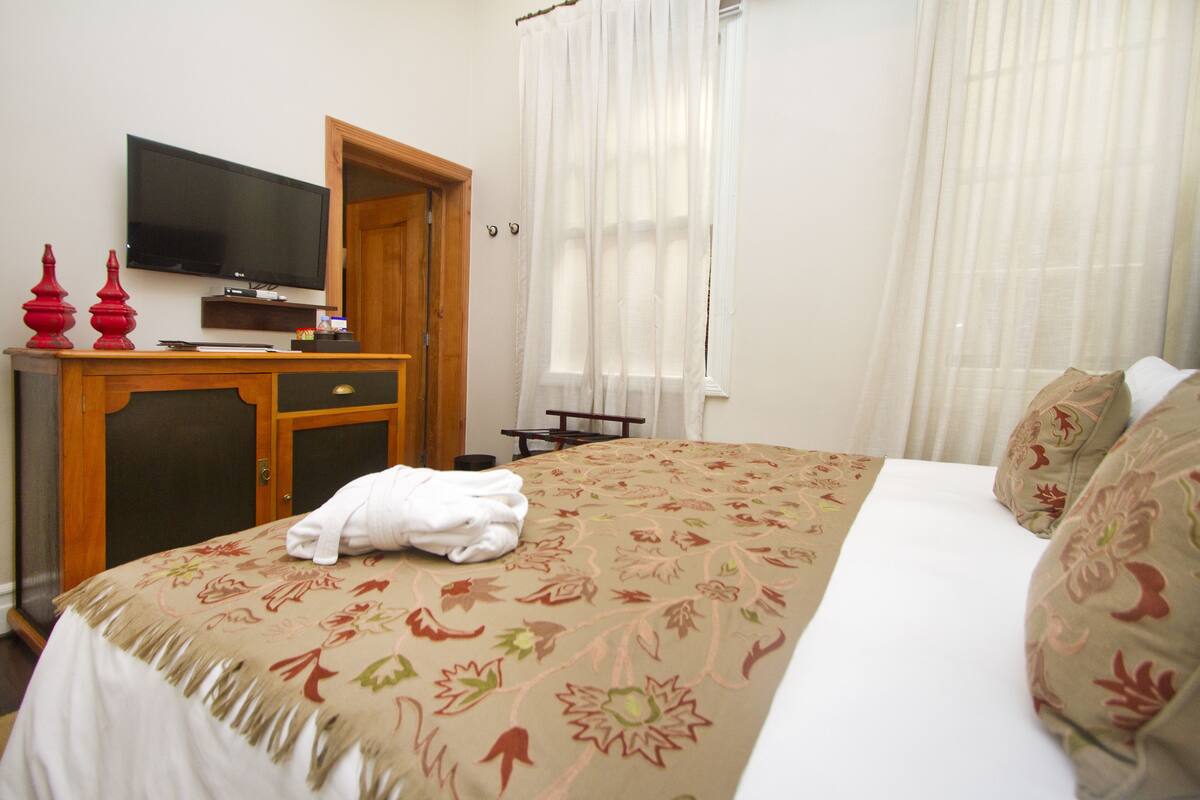 A hotel room with a bed, a television on a wooden stand, a desk with a chair, and a window with curtains.