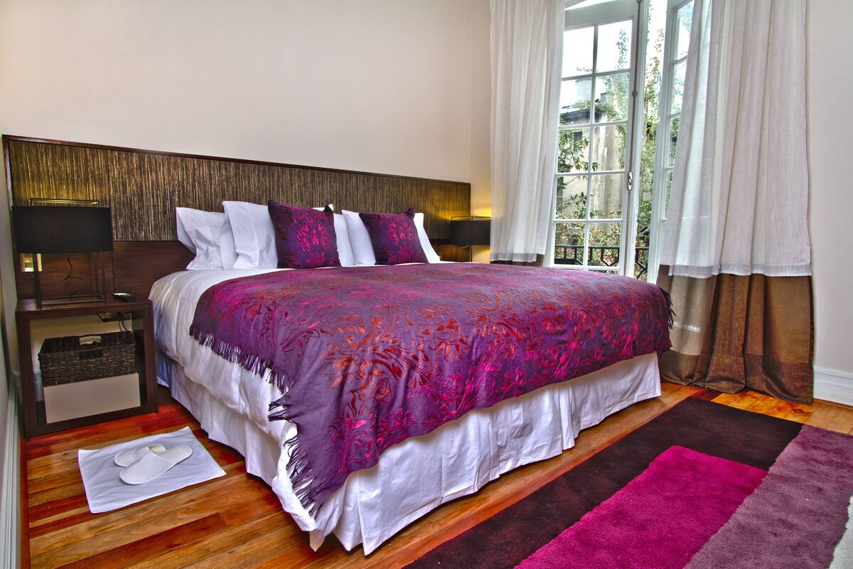 A well-appointed bedroom with a large bed, a nightstand, a lamp, and a view of the outdoors through a window.