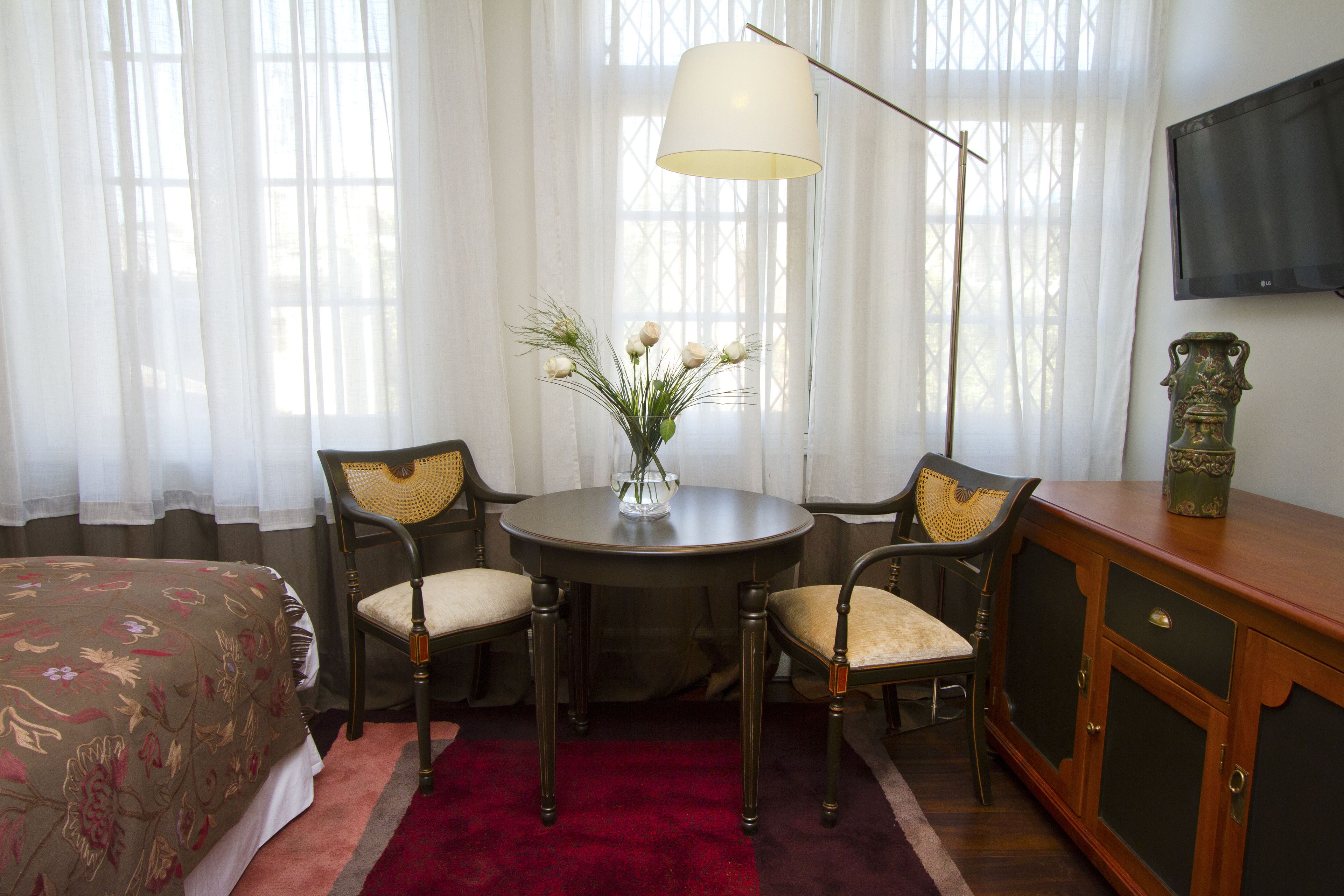 Une chambre d’hôtel avec un lit, une table ronde entourée de chaises, un vase avec des fleurs, une lampe et un téléviseur fixé au mur.