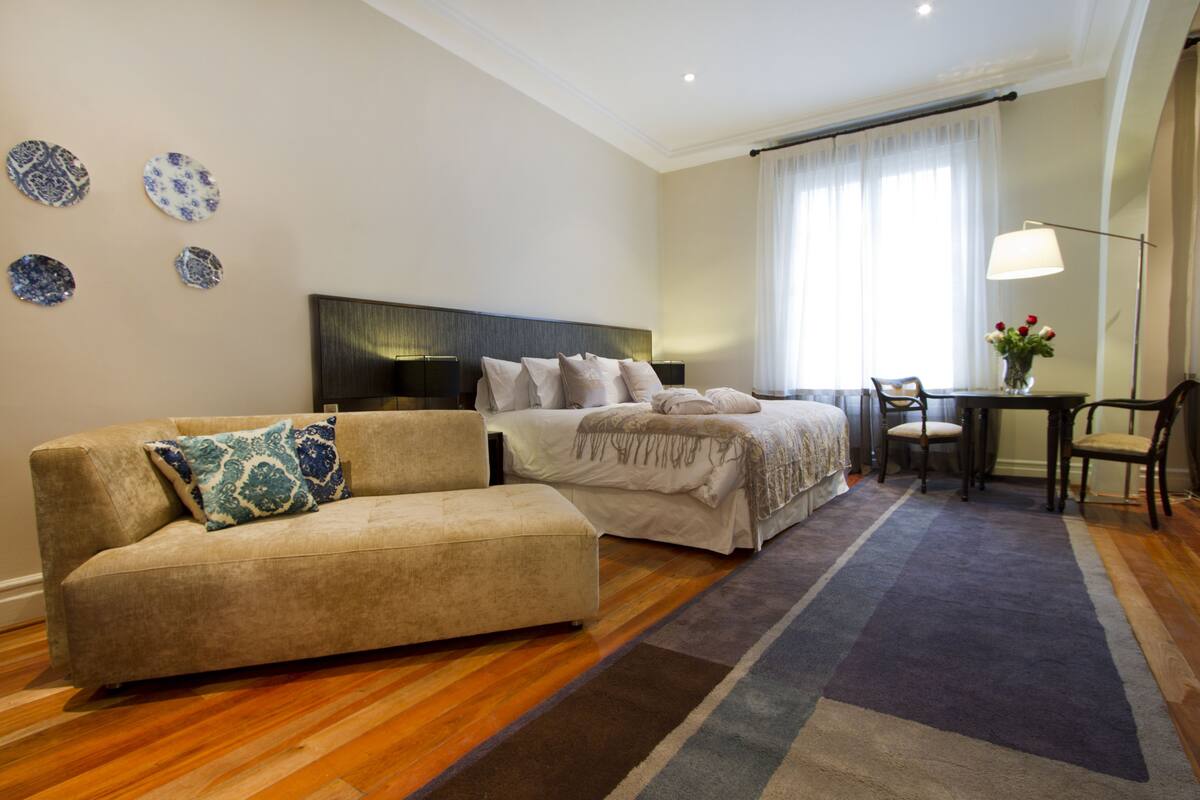 A modern hotel room with a sofa, a bed, a desk with a chair, and a lamp.