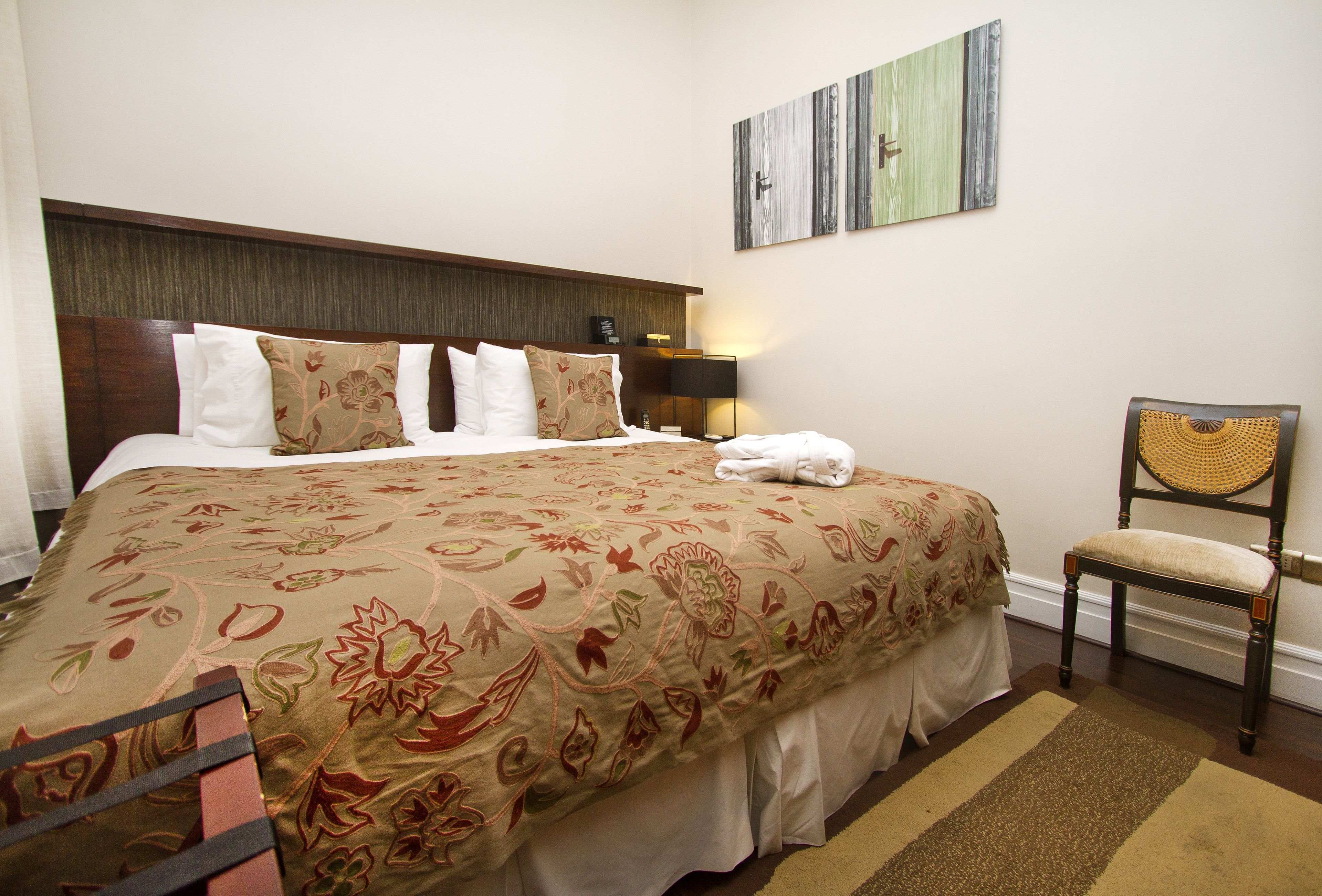 Una cama bien hecha con colcha floral, una lámpara de noche y una silla con respaldo trenzado.