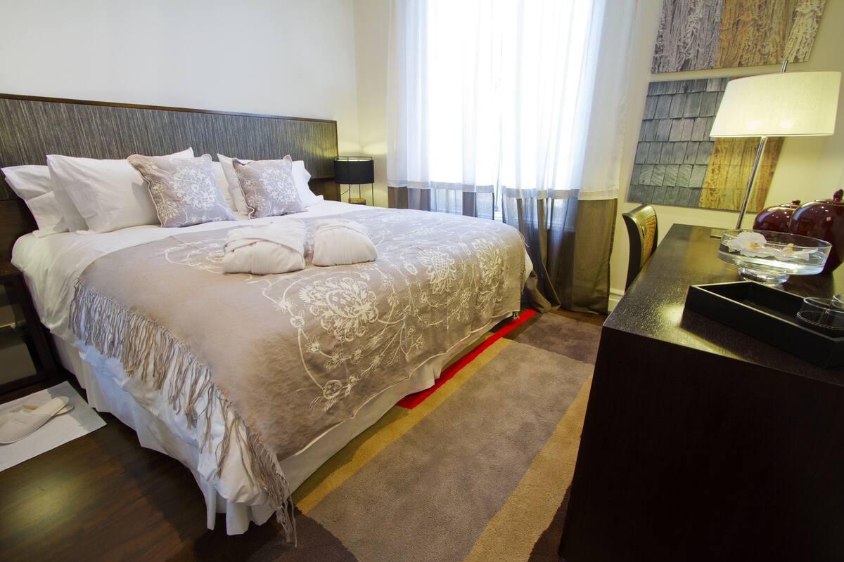 A neatly made bed with a white and beige bedspread, a bedside table with a lamp, and a view of the outside through sheer curtains.