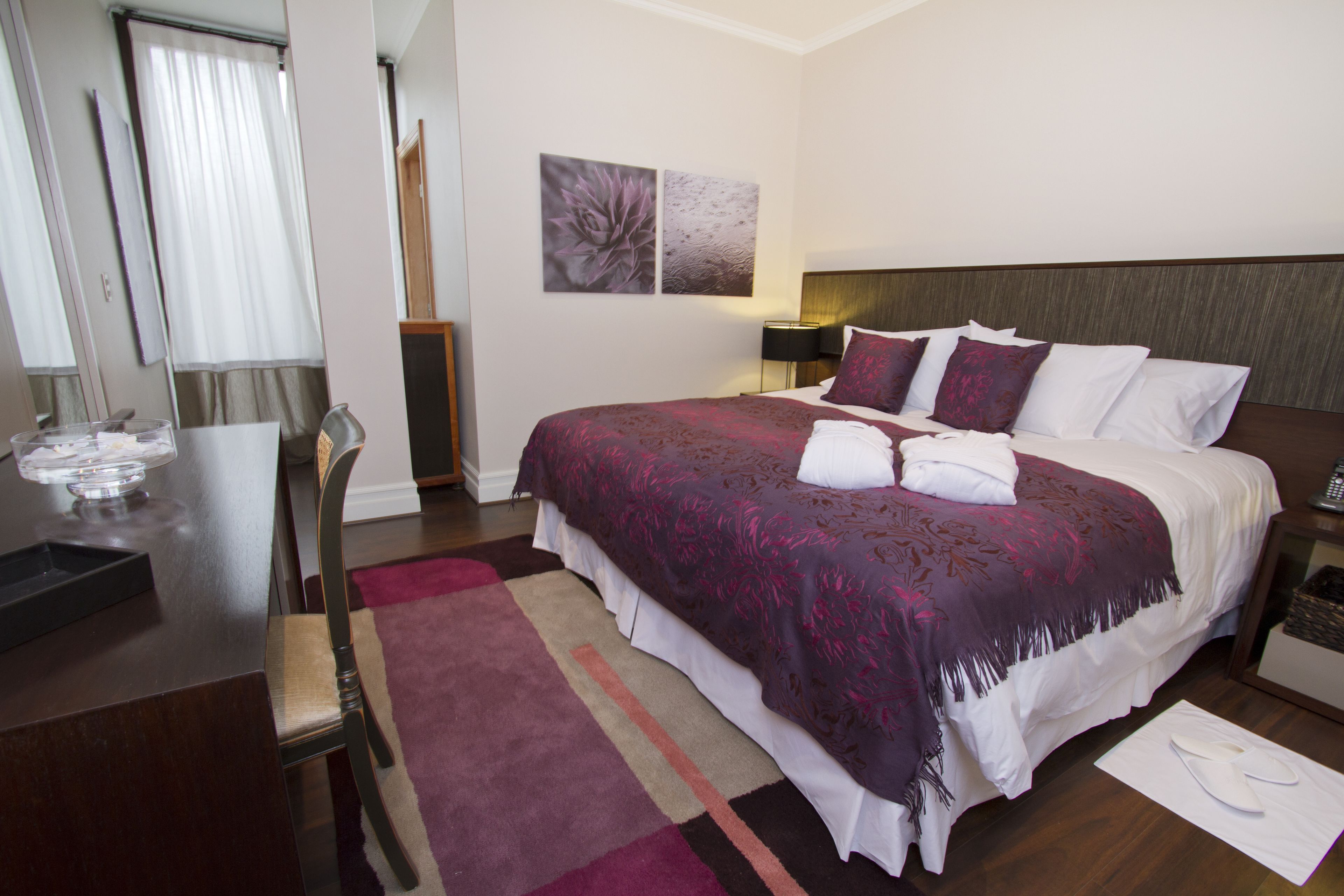 A hotel room with a large bed, a desk with a chair, a nightstand, and a wall-mounted lamp.
