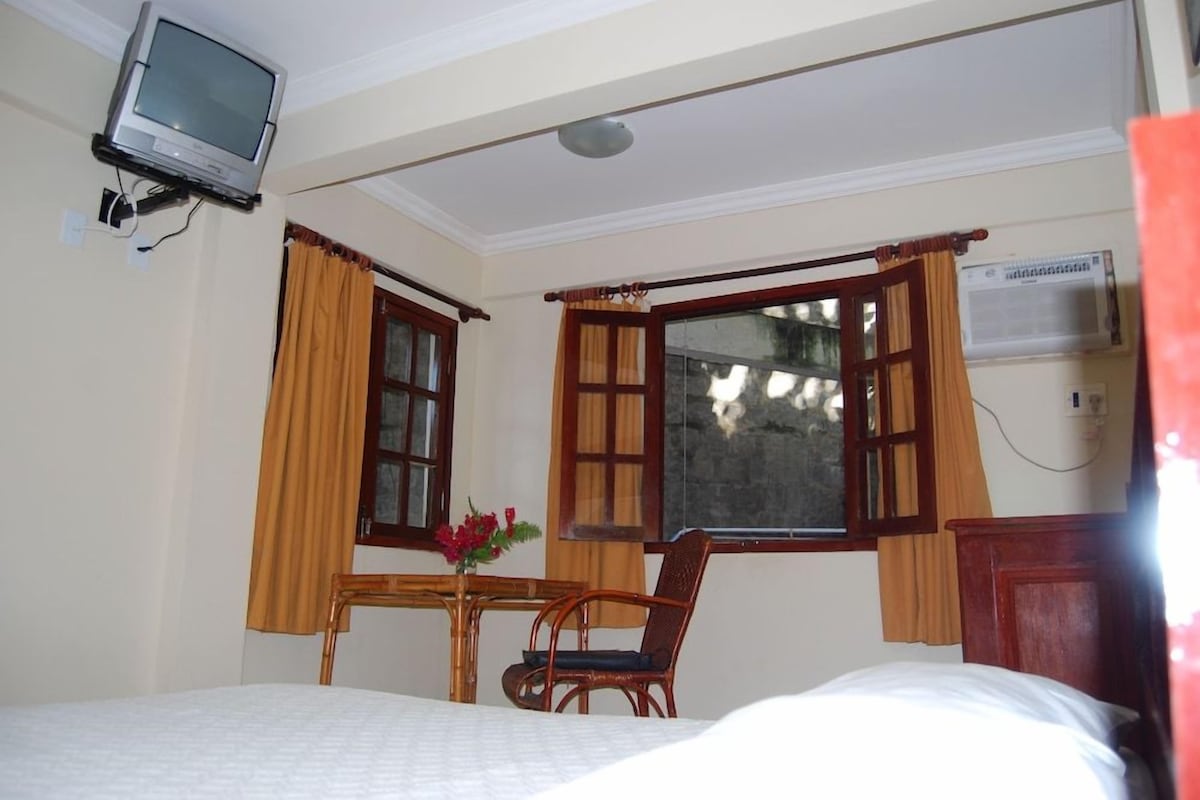 A hotel room with a bed, a desk with a chair, a television mounted on the wall, and a window with curtains.