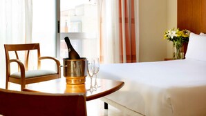 Een hotelkamer met een bed, een houten stoel, een tafel met een fles en glazen, en een vaas met bloemen.