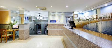 A modern hotel lobby with a reception desk, a seating area, a map on the wall, and a large window with a view of a beach and buildings.