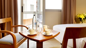 Een hotelkamer met een bed, een houten tafel met een stoel, een vaas met gele bloemen en een raam met gordijnen.