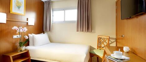 A hotel room with a bed, a TV, a small table with a cup and saucer, a chair, and a window with curtains.