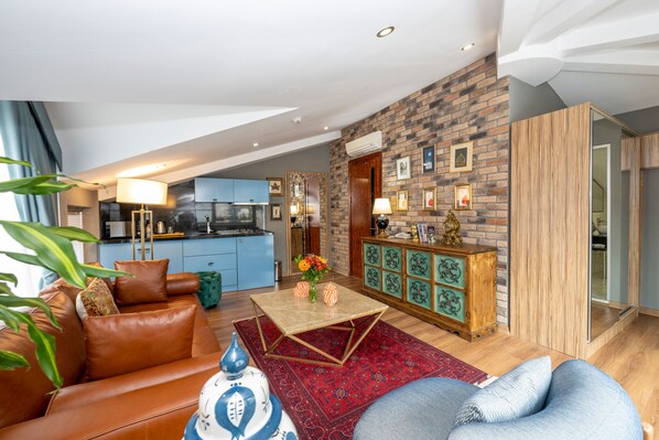 Un salon moderne avec un canapé, une table basse et un tapis rouge. On aperçoit, au fond, une cuisine avec des meubles bleus et un mur en briques.