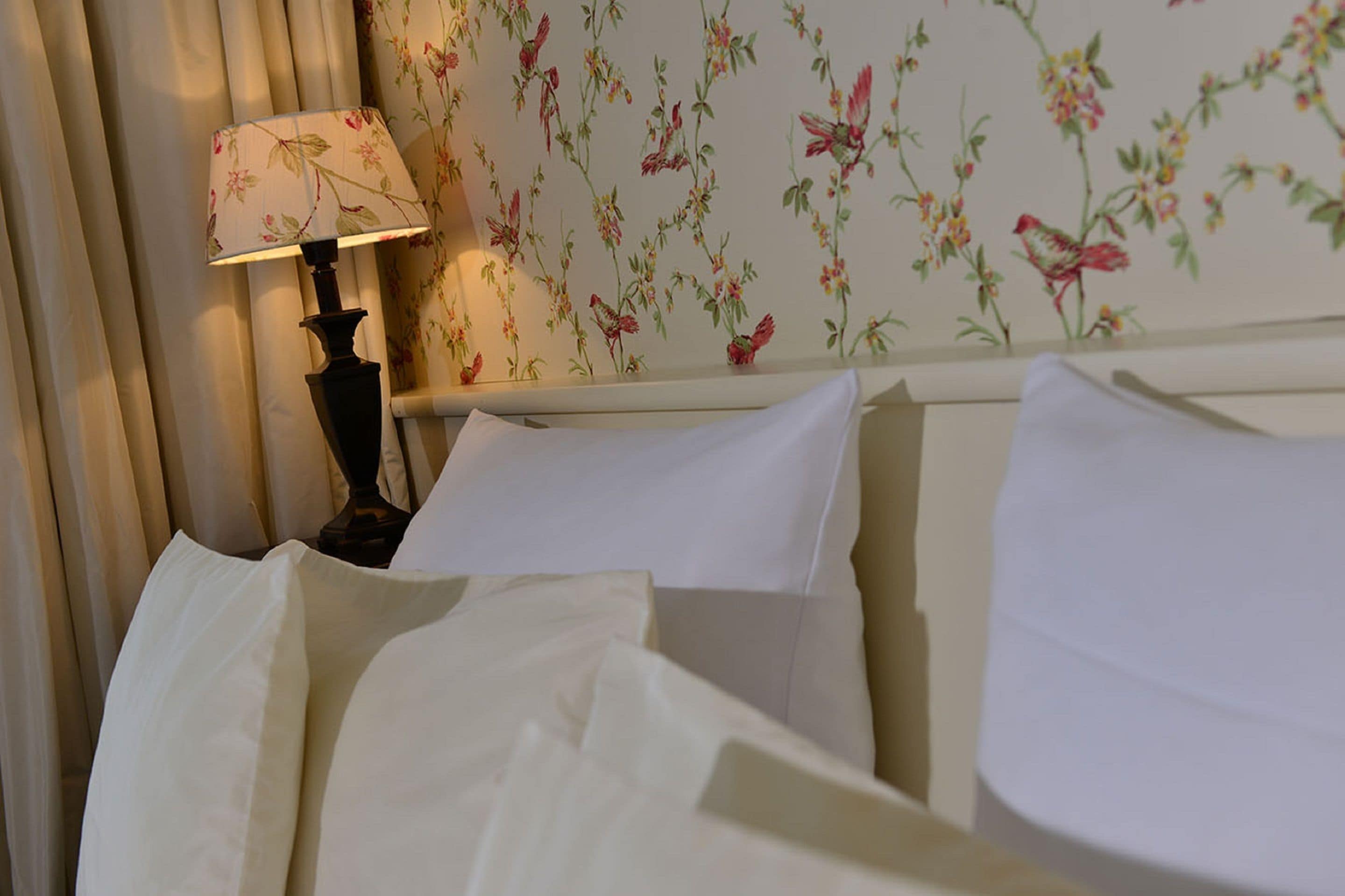 A neatly made bed with white linens and a lamp with a floral lampshade on a bedside table.