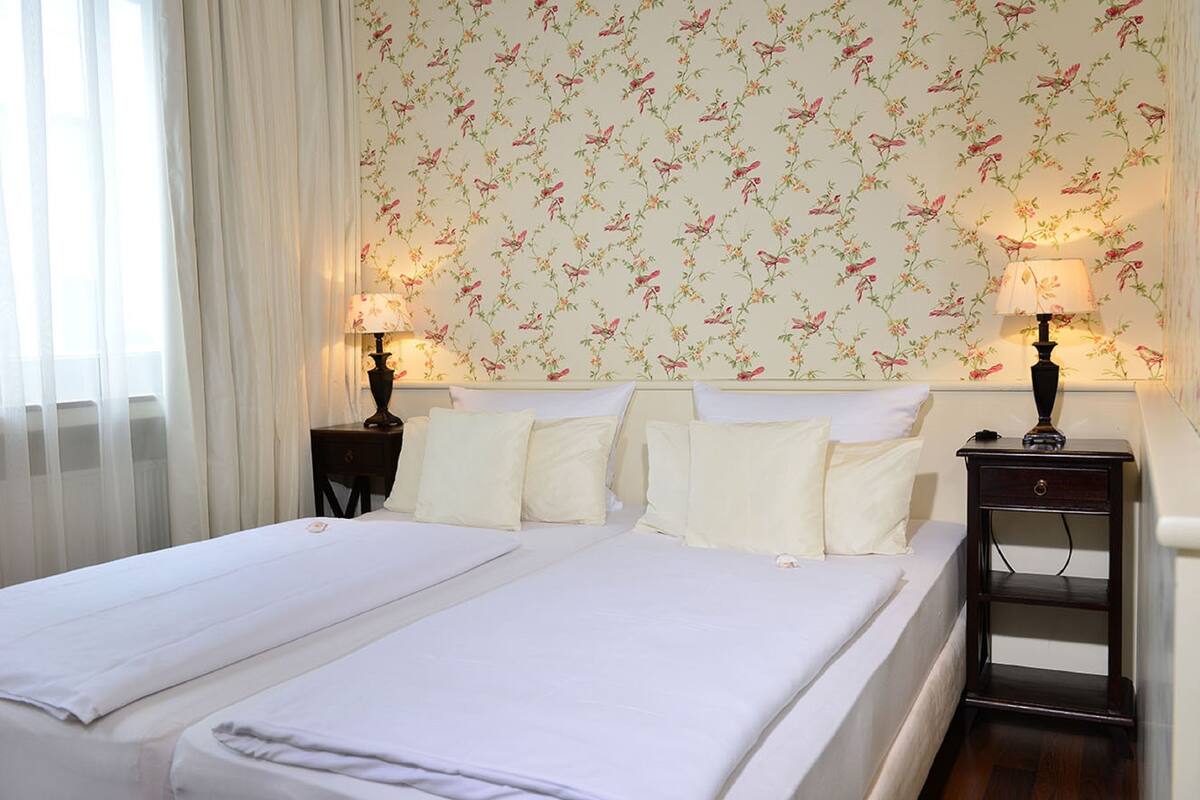 Un lit double avec du linge de lit blanc, deux tables de chevet avec des lampes et un papier peint à motifs floraux.