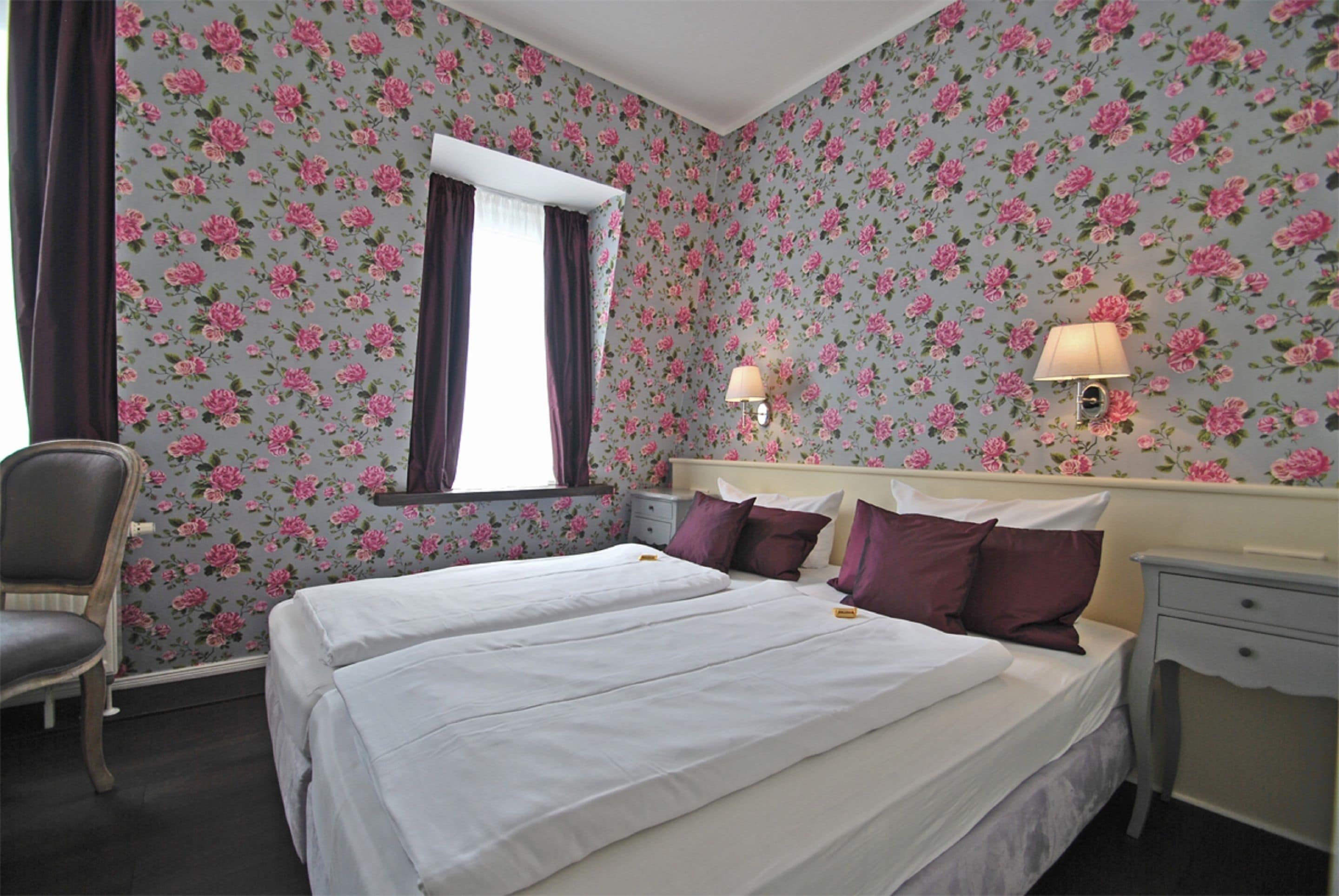 Ein Hotelzimmer mit floralem Wandpapier, zwei Betten, einem Sessel, einem Nachttisch und zwei Wandleuchten.