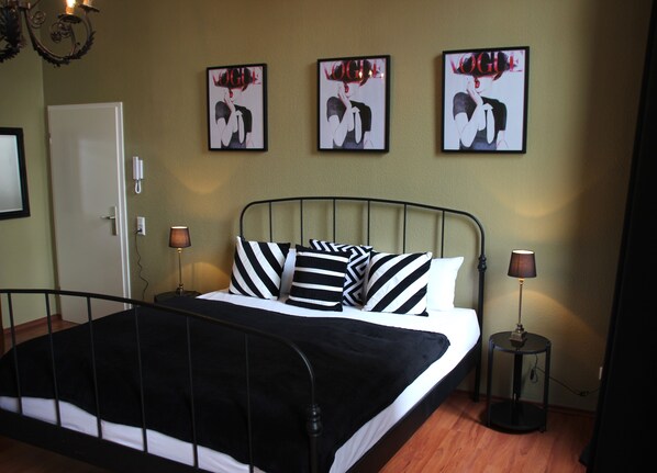 Una cama bien hecha con almohadillas a rayas blancas y negras, un cabecero de metal y una mesita de noche con una lámpara. Tres cuadros enmarcados de una mujer con la palabra "VOGUE" están colgados en la pared sobre la cama.