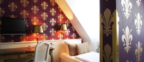 A bedroom with a bed, a desk, and a chair. There is a television on the wall and two lamps on the desk.