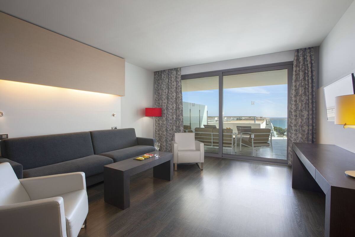 A modern living room with a grey sofa, a coffee table, a white armchair, and a desk. There is a large window with a view of the city and a balcony with outdoor furniture.