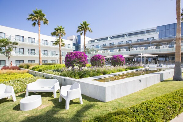 A modern hotel courtyard with white outdoor furniture, a water feature, and lush landscaping including palm trees and purple flowers. - Hipotels Gran Conil (Conil de la Frontera)