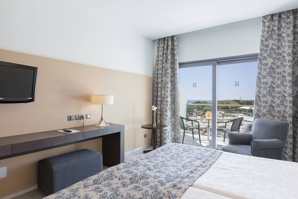 A hotel room with a bed, a desk, a TV, a chair, and a balcony with a table and chairs.