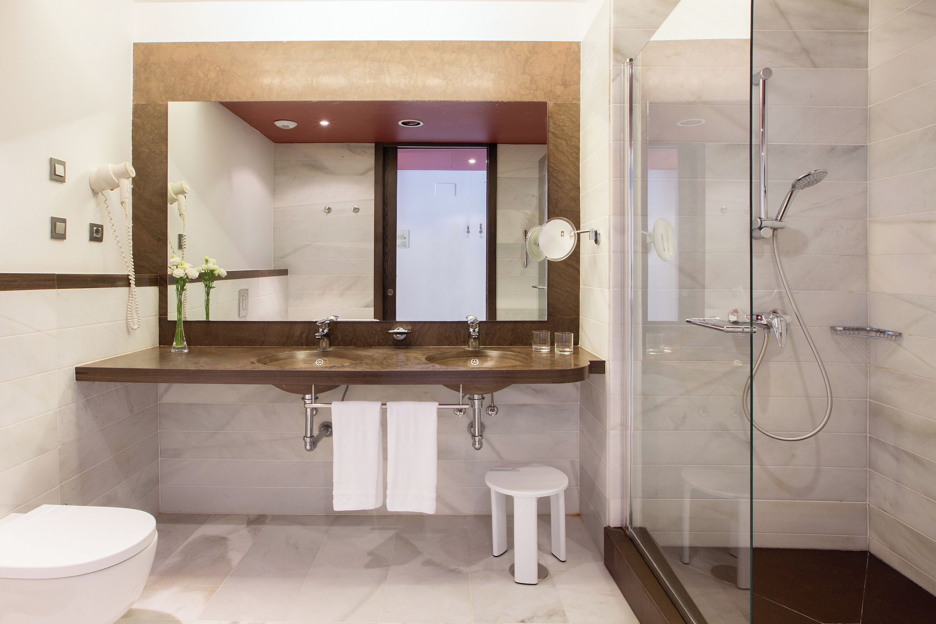 A modern bathroom with a large mirror, two sinks, a toilet, and a glass-enclosed shower.