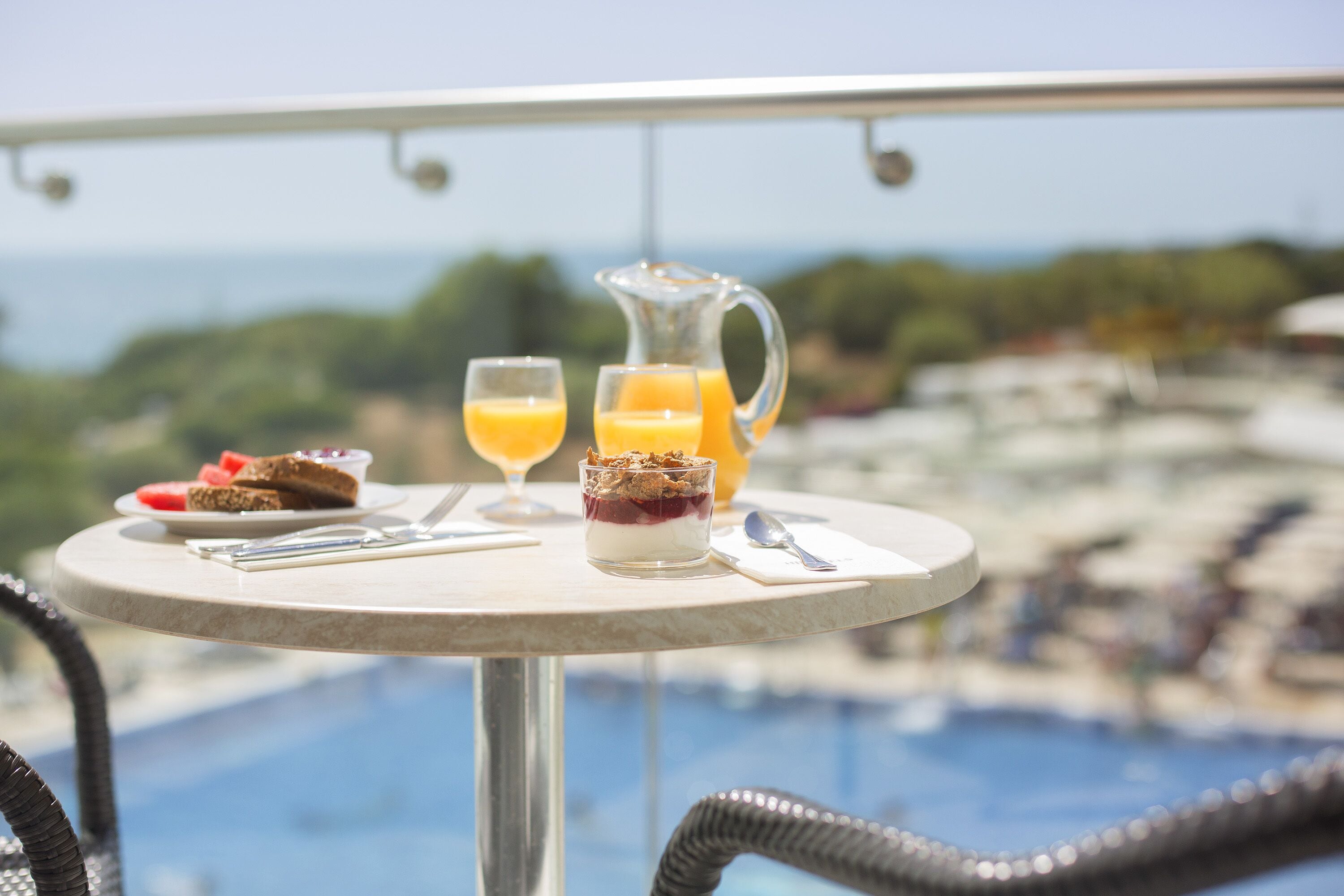 Una mesa con un postre, dos vasos de jugo de naranja y una jarra de jugo, con vista a una piscina y un paisaje.