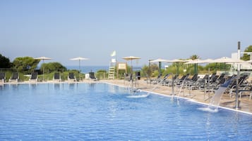 Ein Swimmingpool mit Liegestühlen und Sonnenschirmen, umgeben von Grünpflanzen und einem klaren Himmel.