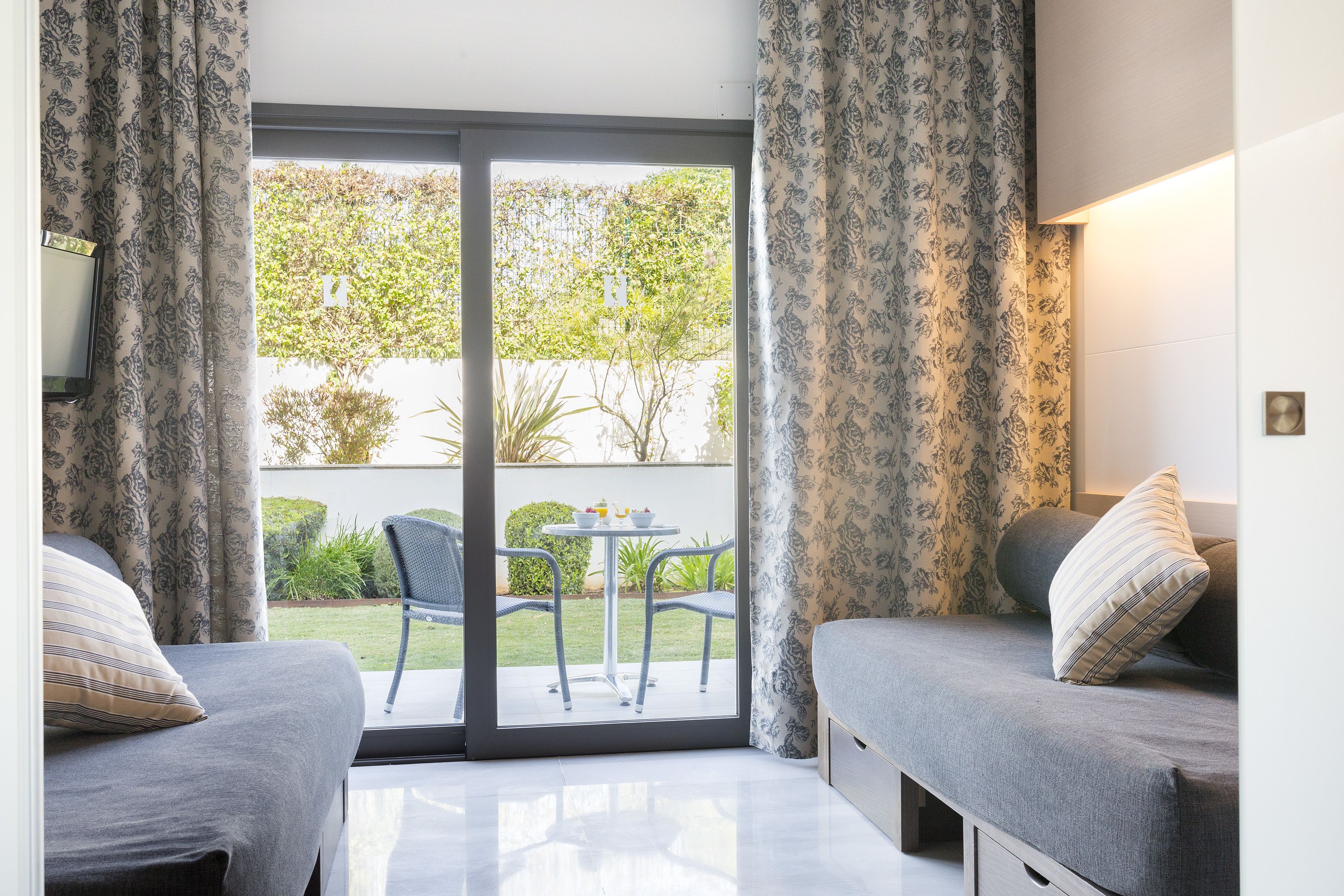A room with two beds, a sliding glass door to a patio with a table and chairs, and a view of greenery outside.