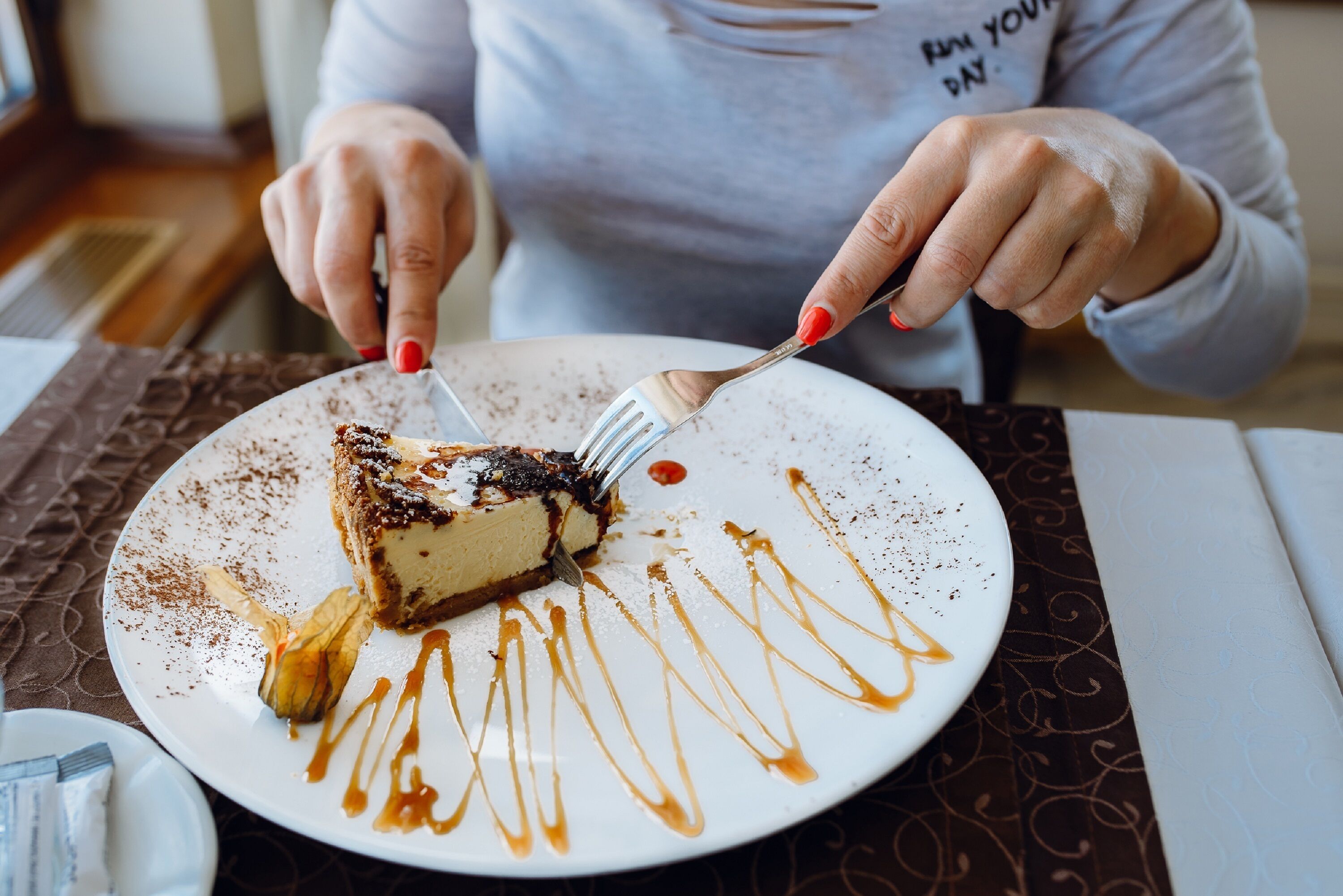 A person cutting a slice of cheesecake with caramel sauce on a white plate.