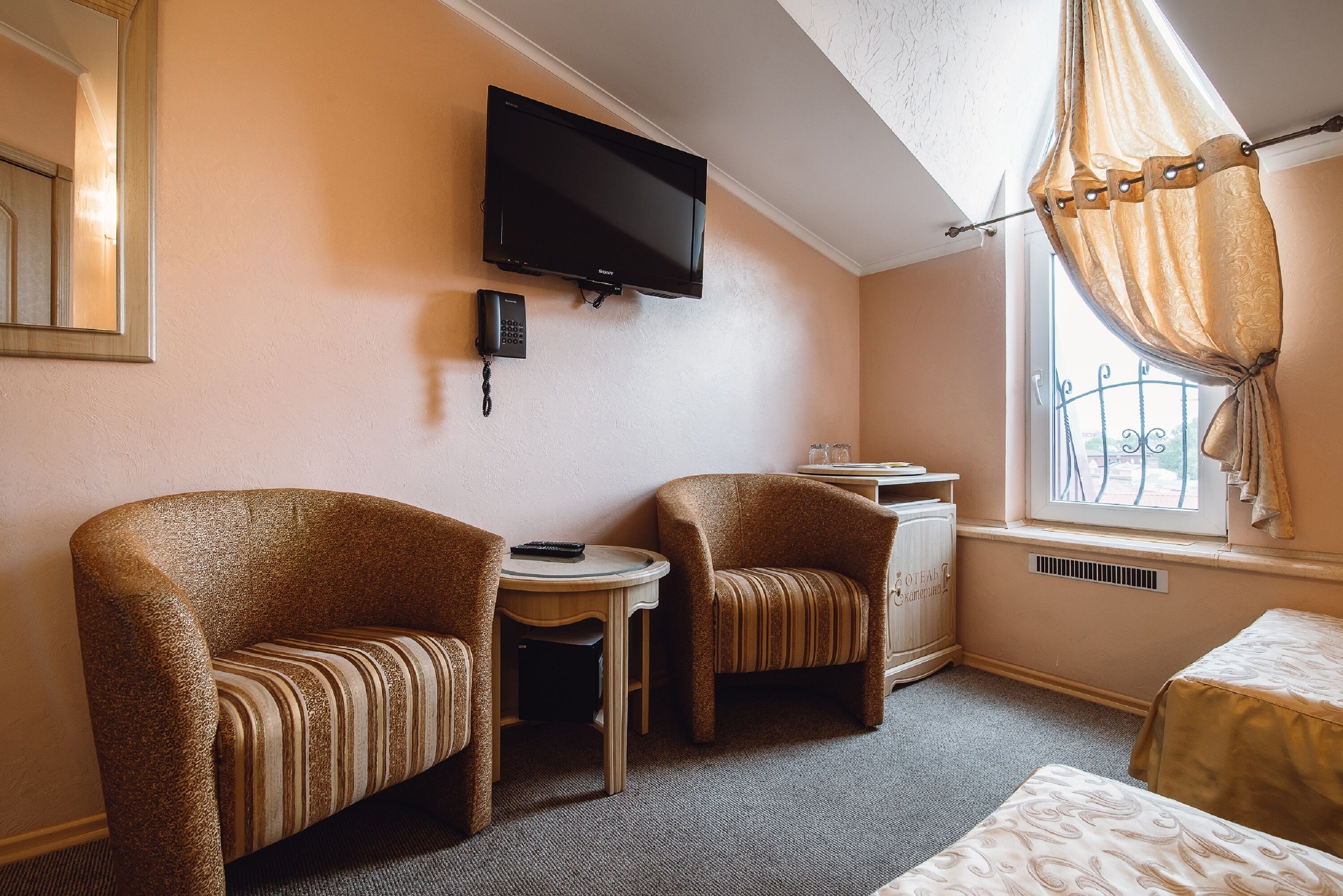 Ett hotellrum med två fåtöljer, ett litet bord, en tv som är monterad på väggen, en telefon och ett fönster med gardiner.
