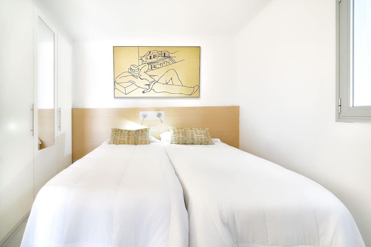 Un lit double avec une literie blanche, une tête de lit en bois et deux coussins à motifs. Un tableau encadré représentant un bâtiment et une personne est accroché au-dessus du lit.