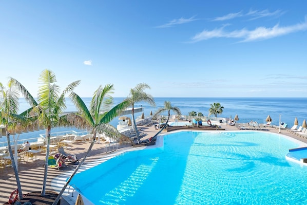 A resort pool area with clear blue water, palm trees, and a view of the ocean.
