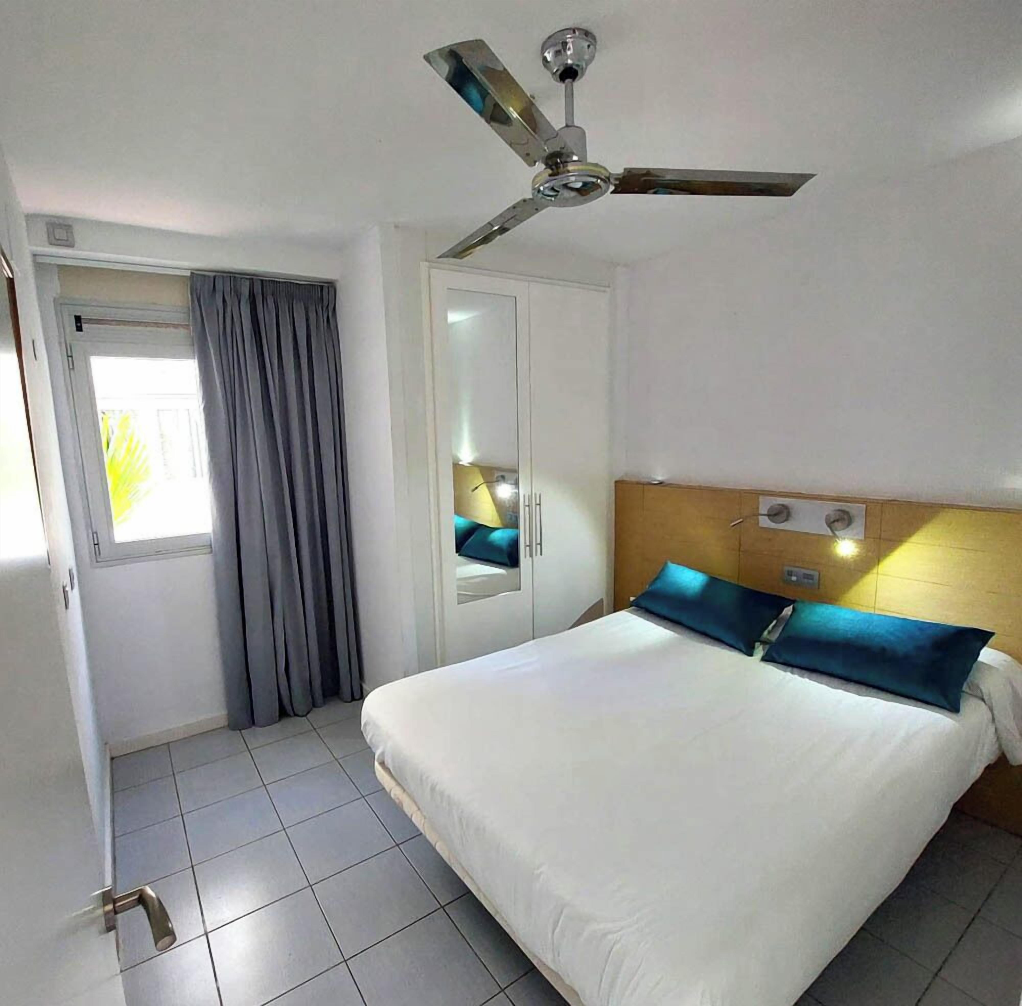 A hotel room with a bed, a ceiling fan, a window with curtains, and a door.