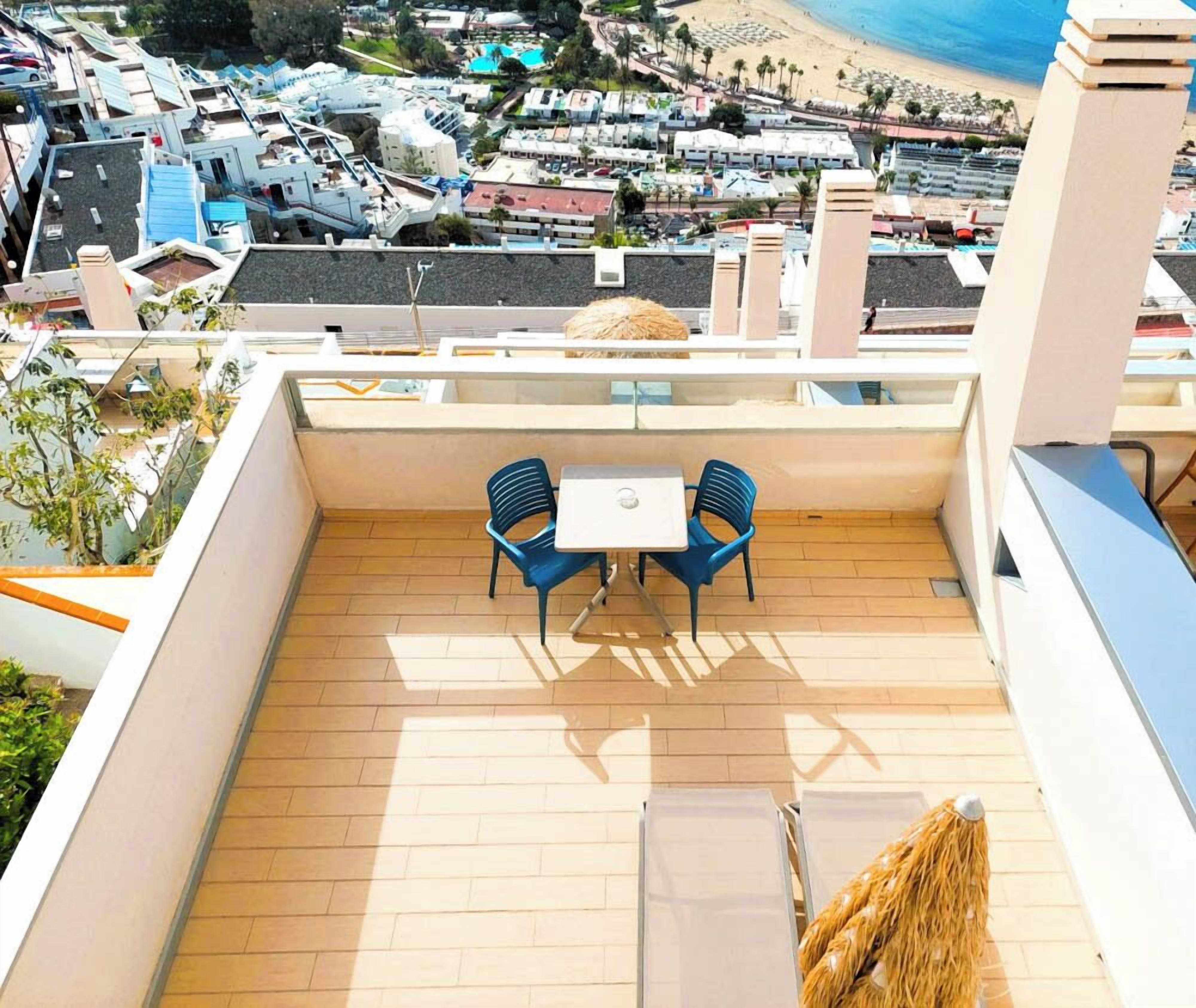 Een terras op het dak met een tafel en twee stoelen, met uitzicht op een strand en een woonwijk.