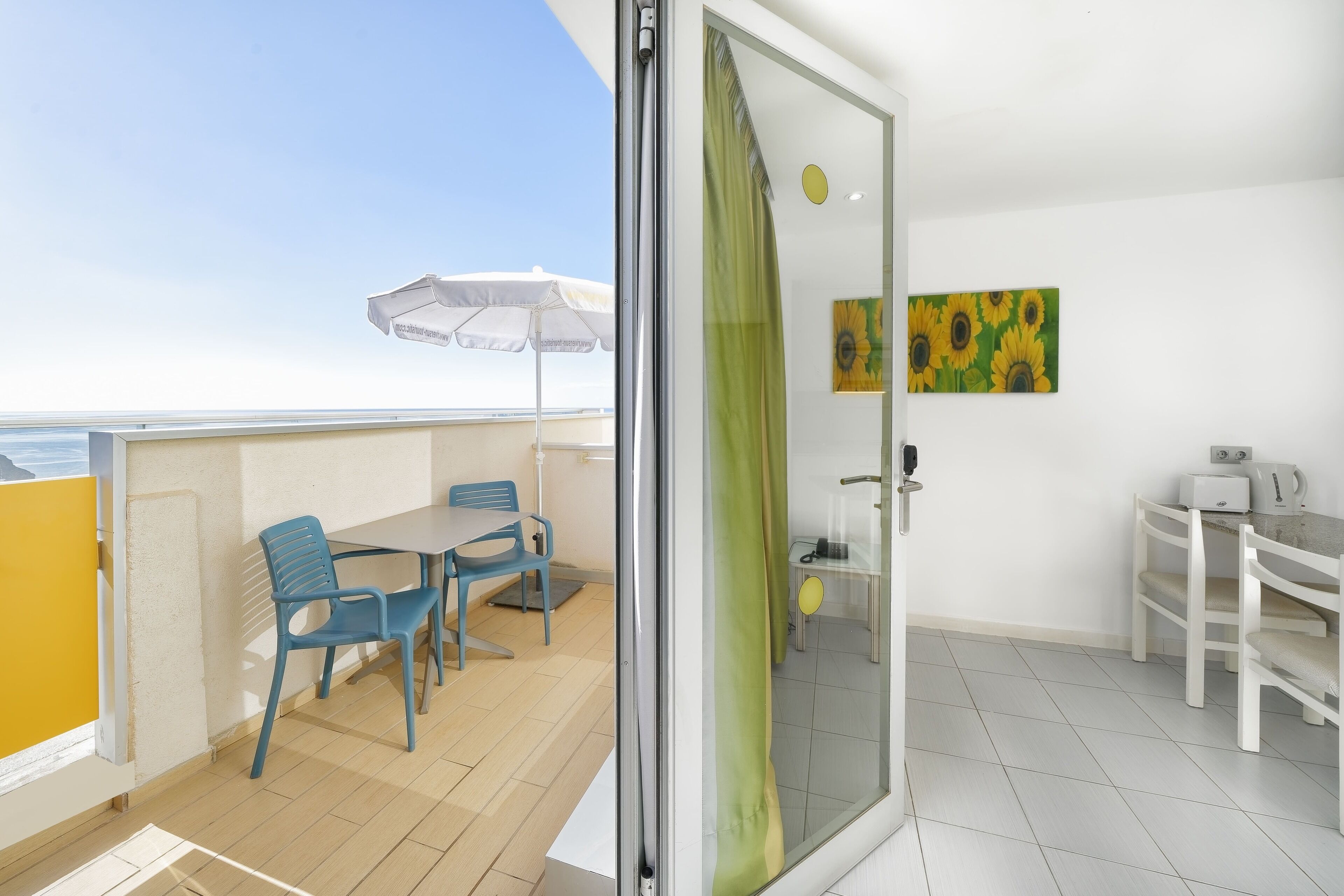 Un balcón con mesa y sillas, una habitación interior blanca con un cuadro, y vista al mar.