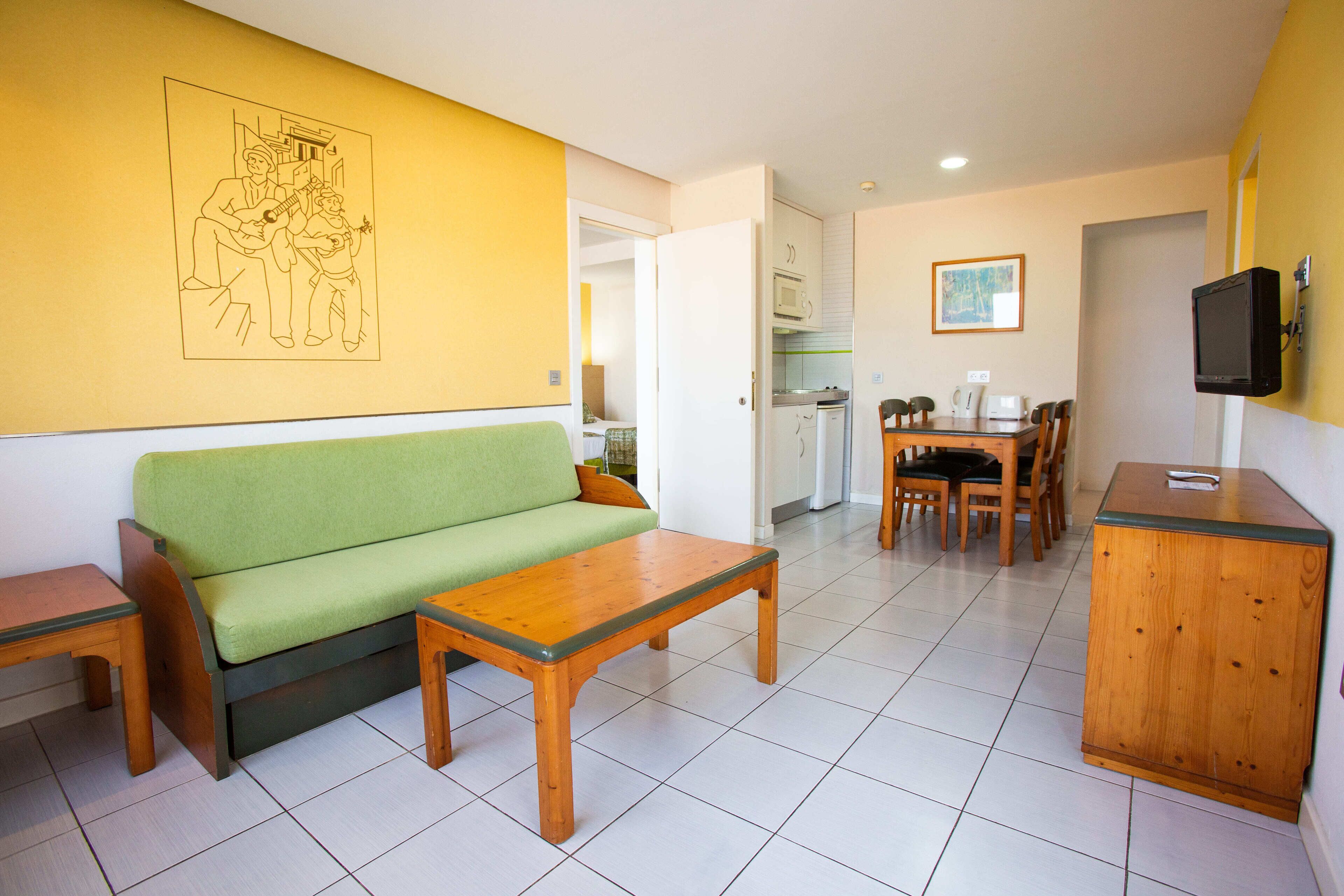 Een kamer met een groene bank, houten salontafel, een eettafel met stoelen, een keukenhoek met een magnetron, en een televisie aan de muur.