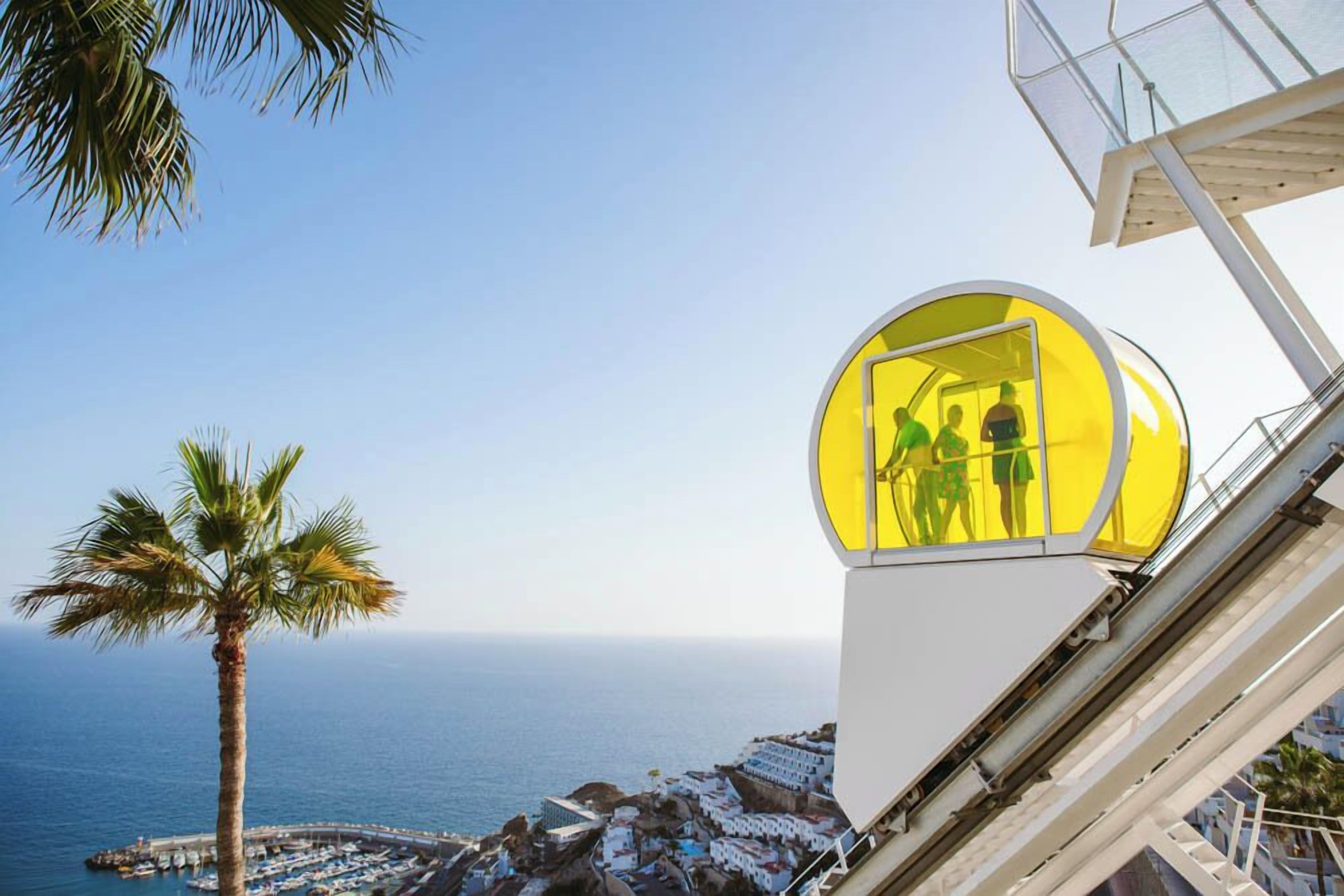 A cable car with passengers, overlooking a coastal town with a palm tree and the sea.