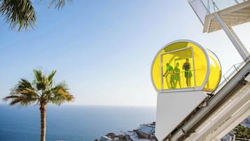 A cable car with passengers, overlooking a coastal town with a palm tree and the sea.