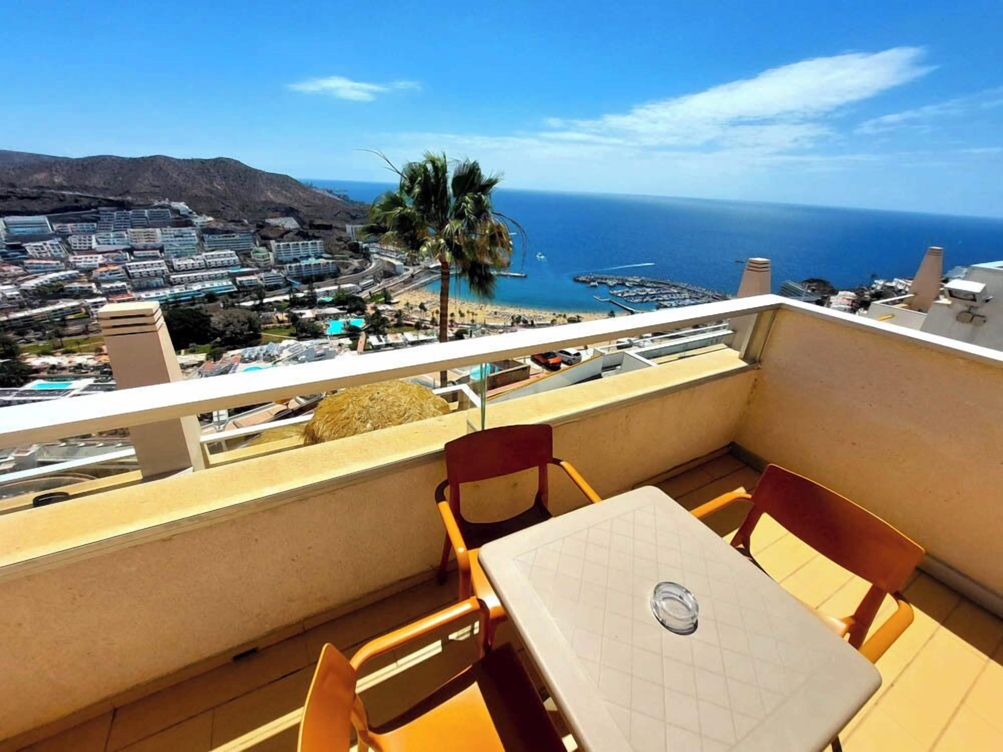 Ein Balkon mit Tisch und Stühlen, der auf eine Küstenstadt mit freiem Blick auf das Meer blickt.