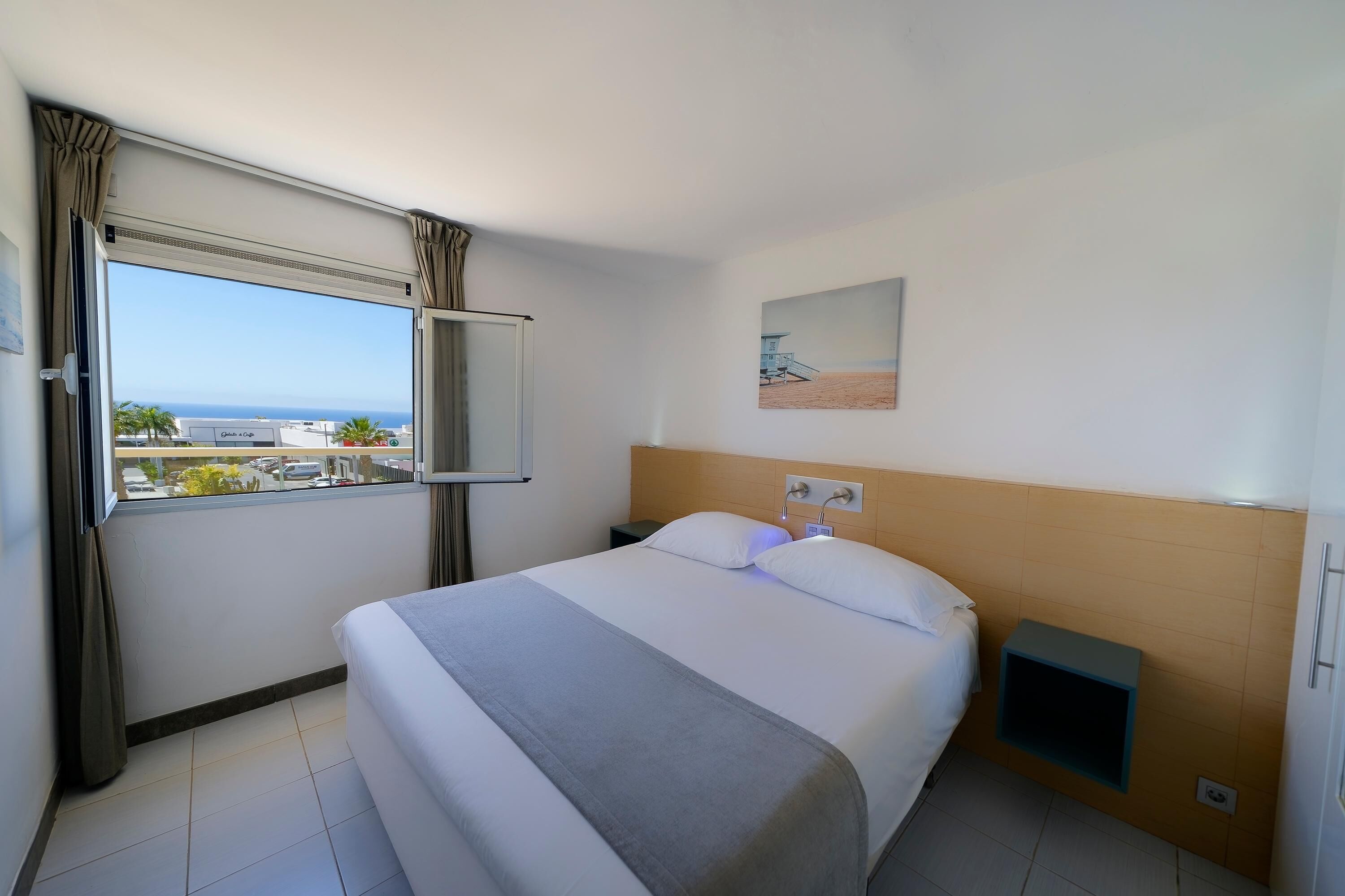 Quarto de hotel com uma cama grande, mesa de cabeceira, televisão na parede, janela com cortinas e um quadro na parede.