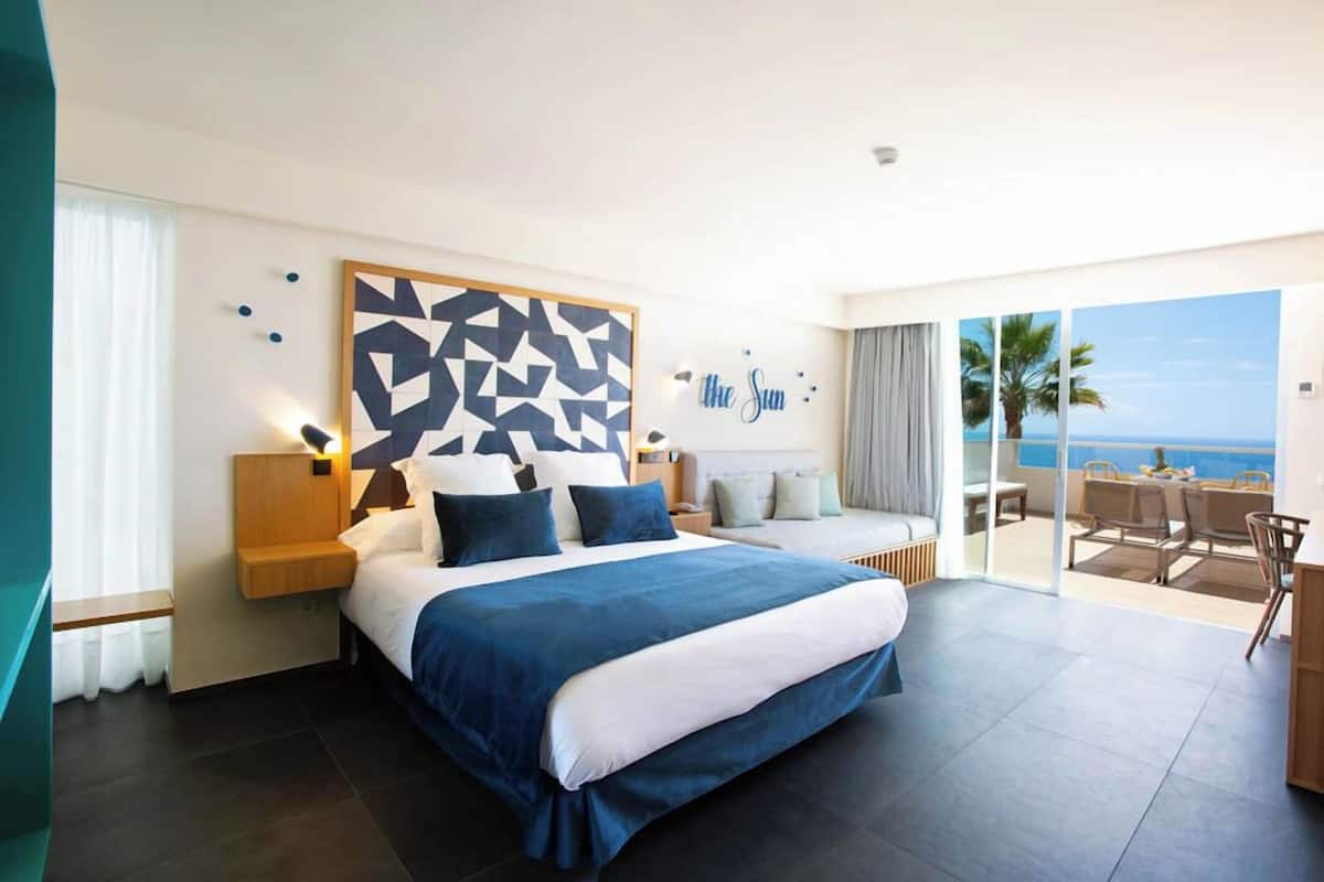 A modern hotel room with a large bed, a geometric wall art piece, a view of the sea, and a balcony with outdoor seating.