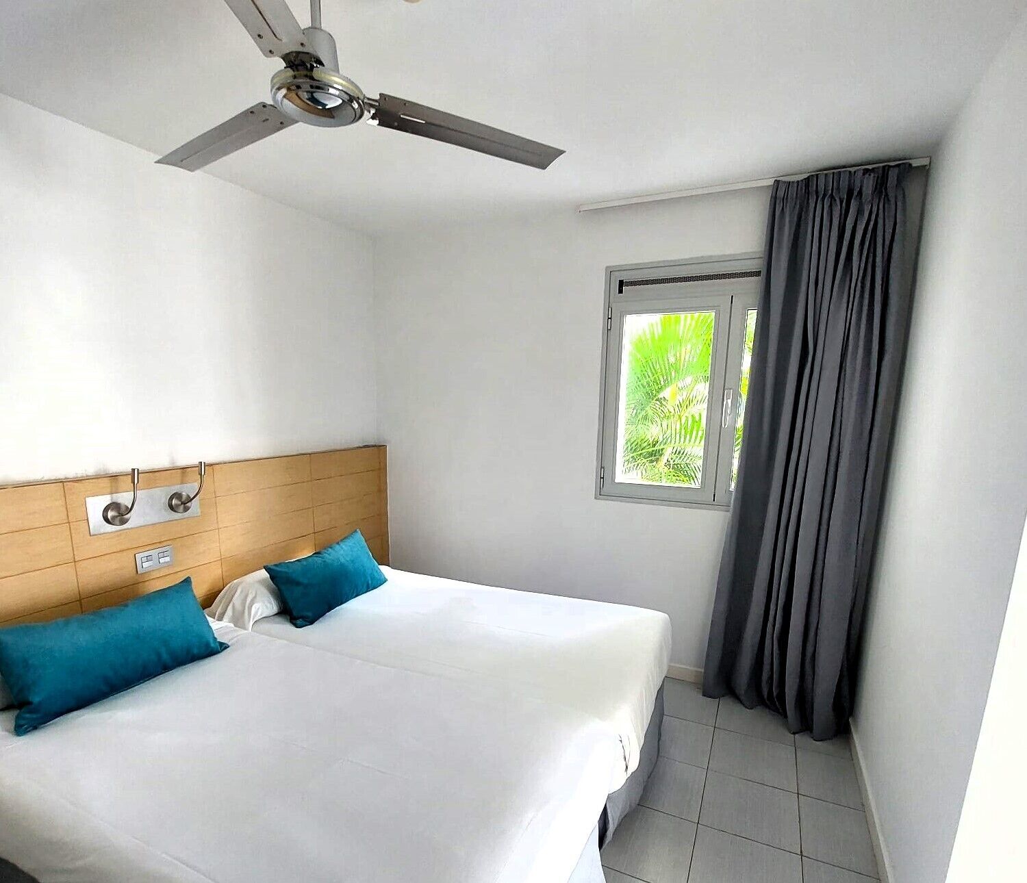 Een hotelkamer met een bed, een plafondventilator, een raam met gordijnen en een aan de muur bevestigde thermostaat.