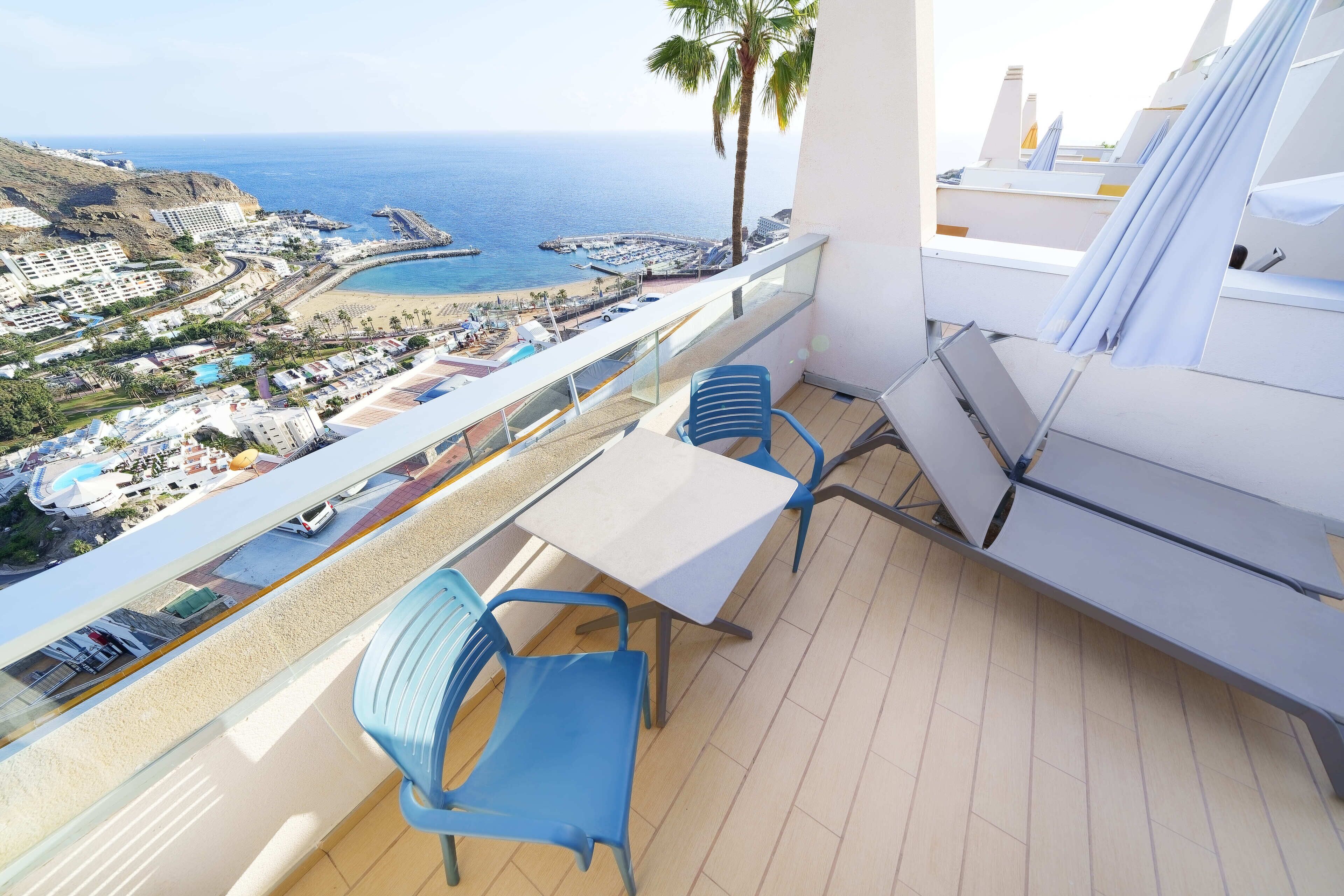 A balcony with a table and chairs, overlooking a coastal area with buildings, a beach, and a pier.