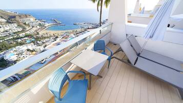 A balcony with a table and chairs, overlooking a coastal area with buildings, a beach, and a pier.